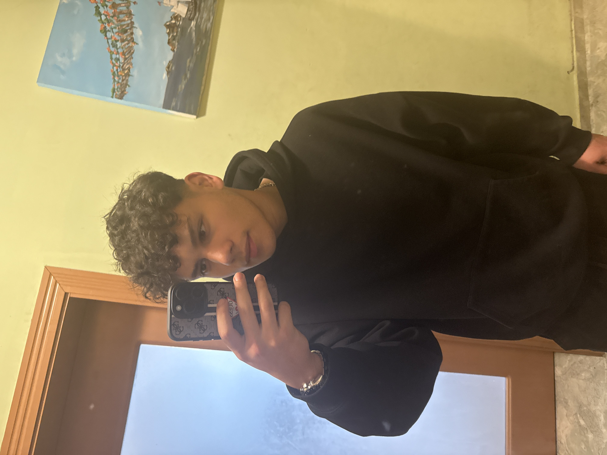 A young man is taking a selfie in what appears to be an indoor setting. He is wearing a black hoodie. He is holding a phone in his left hand. The background shows a light yellow wall with a framed picture hanging on it. Part of a door and a light-colored floor are also visible in the image. The reflection in the door shows a bright sky, suggesting it is daytime. The location cannot be determined to be Sofia, Bulgaria from the visual data provided.