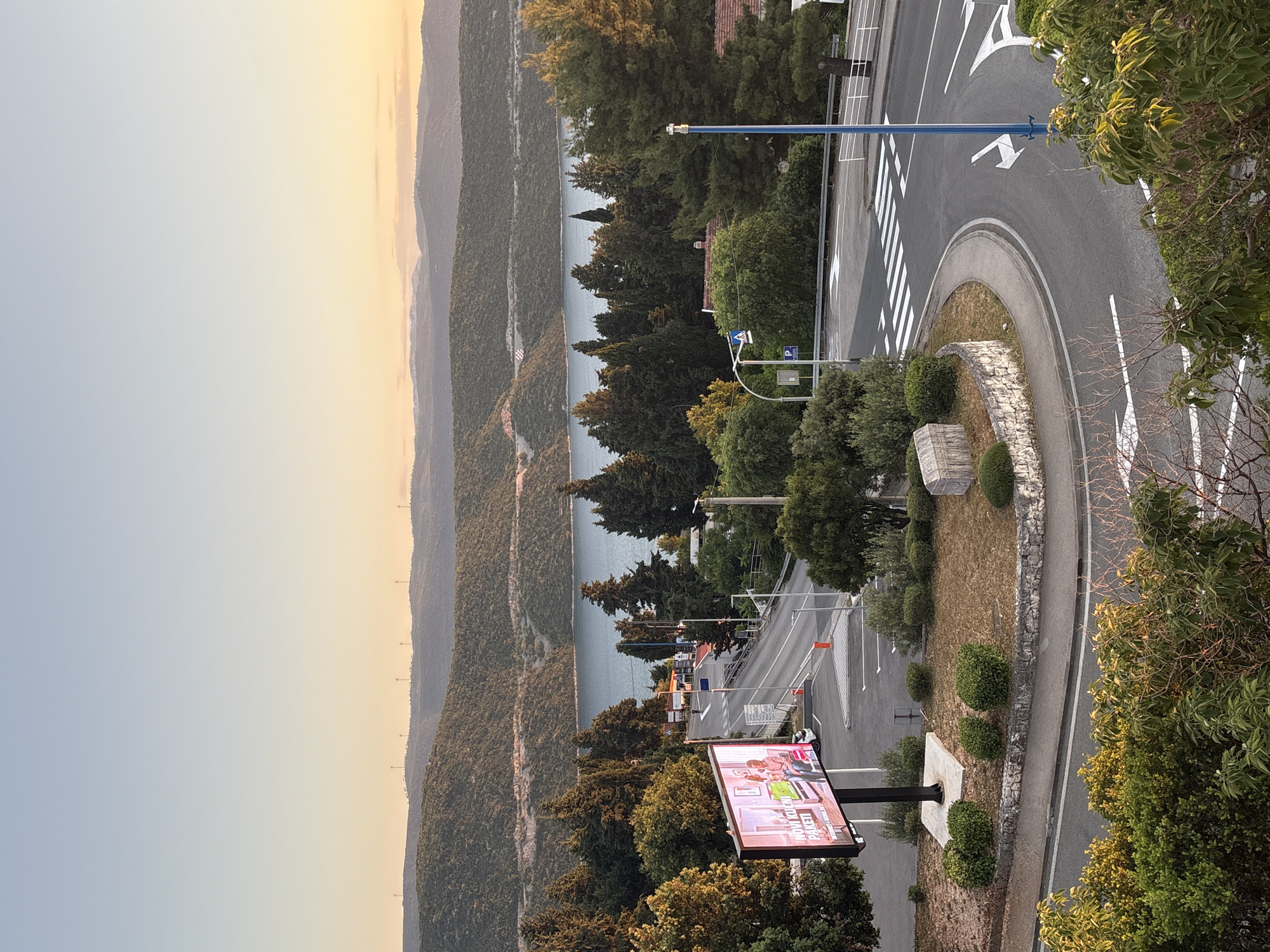 This photograph captures a view of Neum, Bosnia and Herzegovina, from an elevated position, likely a hillside or a high-rise building. The scene includes a landscape view of the mountainous coastline, showcasing hillsides covered in dense vegetation, and a glimpse of the Adriatic Sea. The photograph also features a section of the road network, including a roundabout with white directional arrows and a zebra crossing, suggesting pedestrian access. A digital advertising billboard, showing an ad in Croatian ( "NOVI KUCNI PAKETI"), is positioned among the trees. The photo appears to be taken during the late afternoon or early evening as the sky transitions to light orange.