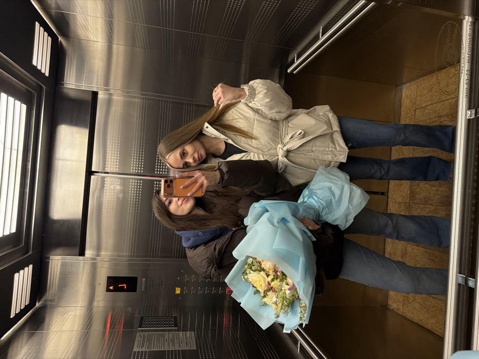 The image captures two women taking a mirror selfie inside an elevator. The photograph itself is rotated 90 degrees clockwise, so the women appear to be standing vertically in the frame, though they are upright within the elevator. Both women are smiling faintly, looking towards the camera.
The woman at the top of the frame has long, straight, light brown hair with blonde highlights. She is wearing a light-colored (possibly off-white or beige) long-sleeved puffer jacket that is tied at the waist, a dark top underneath, and blue jeans. Her left hand is casually placed near her hair.
The woman at the bottom of the frame has long, dark brown hair. She is holding an orange-cased smartphone in her right hand, taking the selfie. She is wearing a dark puffer jacket, likely black or dark brown, over a blue hooded top and blue jeans. In her left arm, she cradles a bouquet of flowers wrapped in light blue paper. The bouquet consists primarily of light-colored flowers, appearing to be white or cream roses, interspersed with some greenery and smaller floral accents.
The elevator interior features metallic walls, likely stainless steel, which reflect the women and the lighting. One wall has a subtle pattern of horizontal lines and small dots. Another wall, to the left, has a window-like panel with vertical slats, allowing some light into the space. An elevator control panel is visible on a metallic wall, displaying a red illuminated digit "1", indicating the current floor, along with a grid of unlit buttons and some indistinct text. Metal handrails are visible on the side walls. The floor of the elevator appears to be a light brown, possibly tiled or patterned. On the right side of the elevator, near a vertical metal bar, there is some text etched or printed onto the wall, which includes "www.2rb.ru", a phone number "990-54-73", and the Russian words "НАГОРНАЯ 17" (NAGORNAYA 17), suggesting a location or address.
The overall mood appears cheerful and pleasant, possibly capturing a moment before or after a special occasion, indicated by the flowers.