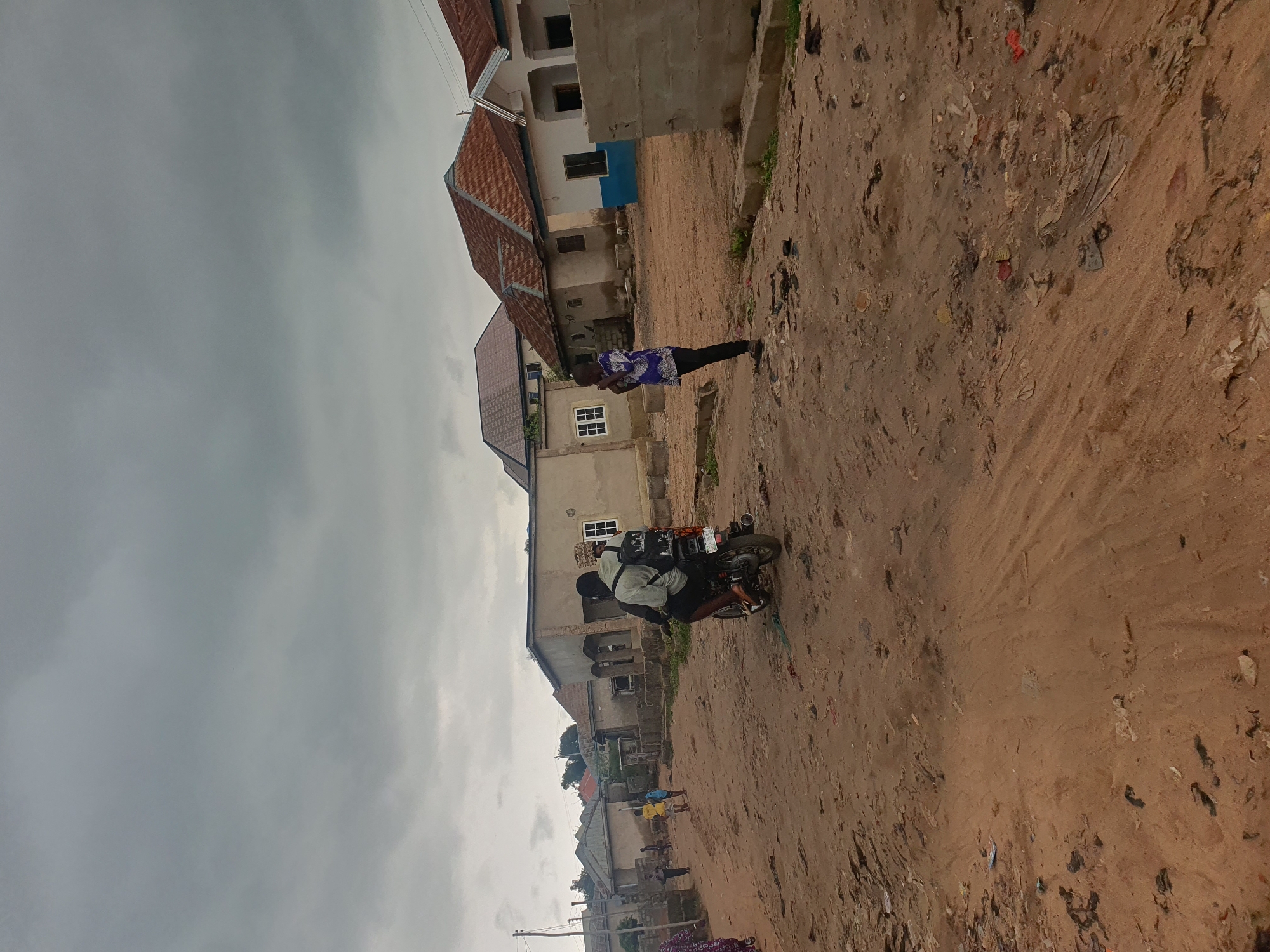 The image depicts a street scene in Lokoja, Nigeria, characterized by a dirt road and several houses under a cloudy sky. In the foreground, a motorcycle with two occupants rides away from the camera. The rider is wearing a light-colored shirt and dark shorts, with a backpack. The passenger is wearing a white shirt, dark pants, and a patterned headwrap. To the right of the motorcycle, a young man stands facing away from the camera, wearing a blue and white patterned shirt and black pants. He appears to be holding his hands in front of him. In the background, a row of houses with corrugated iron roofs lines the street. Some houses appear to be under construction or are unfinished. The ground is covered in sand and debris, suggesting a rural or semi-urban environment. The overall atmosphere is overcast, with a diffused light.