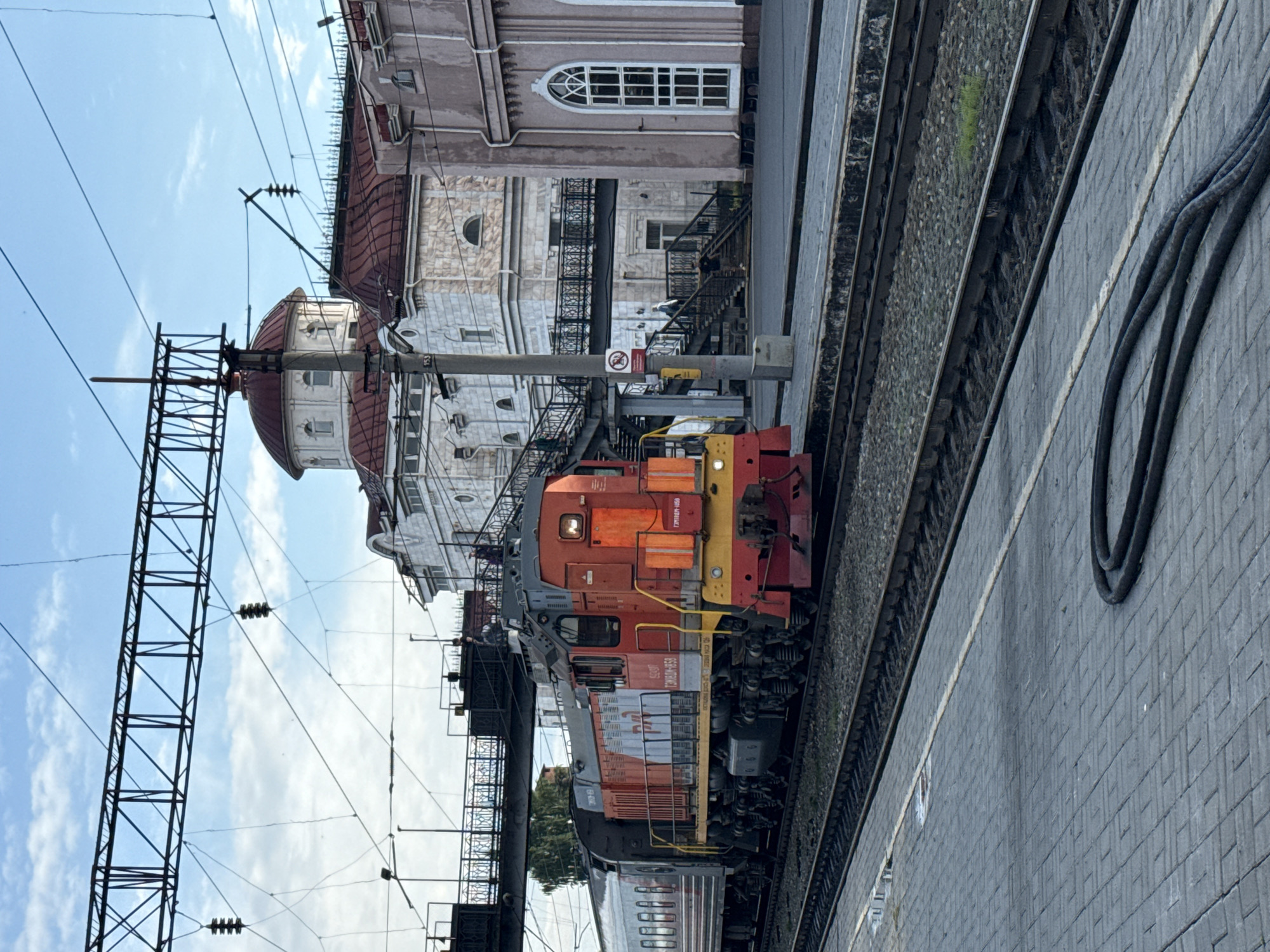 A train with an orange and red locomotive and passenger cars is stopped at a station in Izhevsk, Russia. The locomotive is on the right side of the image, and the passenger cars are on the left. The train is on a track that runs parallel to a paved platform. In the background, there are several buildings, including a white building with a red dome, and a pink building with a large arched window. There are also overhead wires and a pedestrian bridge. Several people are visible on the platform and on the bridge. A black hose is coiled on the platform in the foreground. The sky is blue with some clouds.