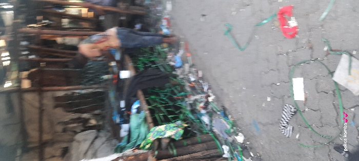 A person is lying motionless amidst debris and scrap materials. The individual, wearing dark pants, is positioned next to what appears to be a makeshift wooden structure. Scattered around are green ribbons, clothing, and general litter, some on the ground. "police kombo" is written in the lower right. The scene is cluttered and chaotic.