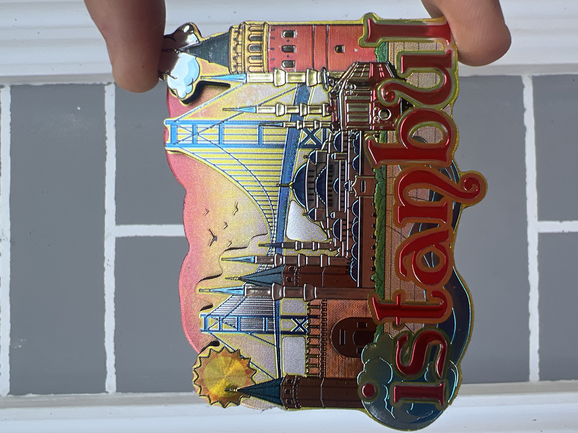 A close-up, eye-level shot shows a magnet with the word "Istanbul" on it. The magnet depicts a cityscape with a large suspension bridge, minarets, domes, and the Galata Tower. The background features a sunset with birds flying. The magnet is held by a person's thumb and index finger, obscuring part of the right side of the magnet. The magnet is positioned against a gray brick wall with white grout. The colors are vibrant and metallic.