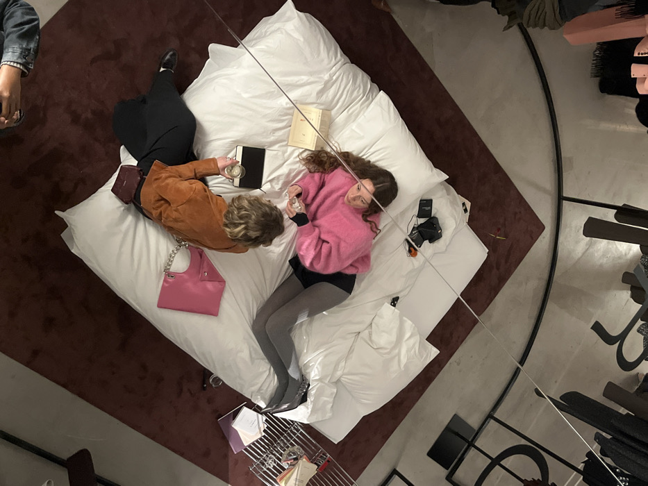 A high-angle photograph, potentially captured via a reflective ceiling surface, depicts a scene within a high-end fashion boutique in Paris, France. Two women are reclining on a large, white, multi-cushioned mattress or daybed centrally positioned on a deep burgundy carpet.
The first woman, located on the upper right, is seen from the chest up, lying on her left side. She wears a brown suede jacket and her short, curly blonde hair is visible. Her left hand holds a clear glass containing liquid. A small burgundy shoulder bag rests behind her, and a black book or tablet is placed on the mattress near her hand.
The second woman, positioned on the lower left, lies on her back with her head slightly elevated by pillows. She wears a bright pink textured sweater, a black skirt, and grey opaque tights paired with dark, pointed-toe heels. Her long, wavy brown hair spreads around her head. She faces the viewer with a slight smile, holding a small, clear object in her right hand. Adjacent to her head, a light-colored box and two small black electronic devices are visible on the mattress.
Between the two women, a bright pink handbag with a silver chain strap is placed on the white surface. In the upper right corner, partially visible hands and forearms of a third individual, clad in a denim jacket, are observed holding a dark-colored smartphone, angled as if capturing the scene.
The lower portion of the image displays a reflection of the surrounding retail space, separated by a thin horizontal line. This reflection reveals sections of clothing racks holding dark garments, stylized black display fixtures, and a light grey floor, consistent with a contemporary luxury retail setting.