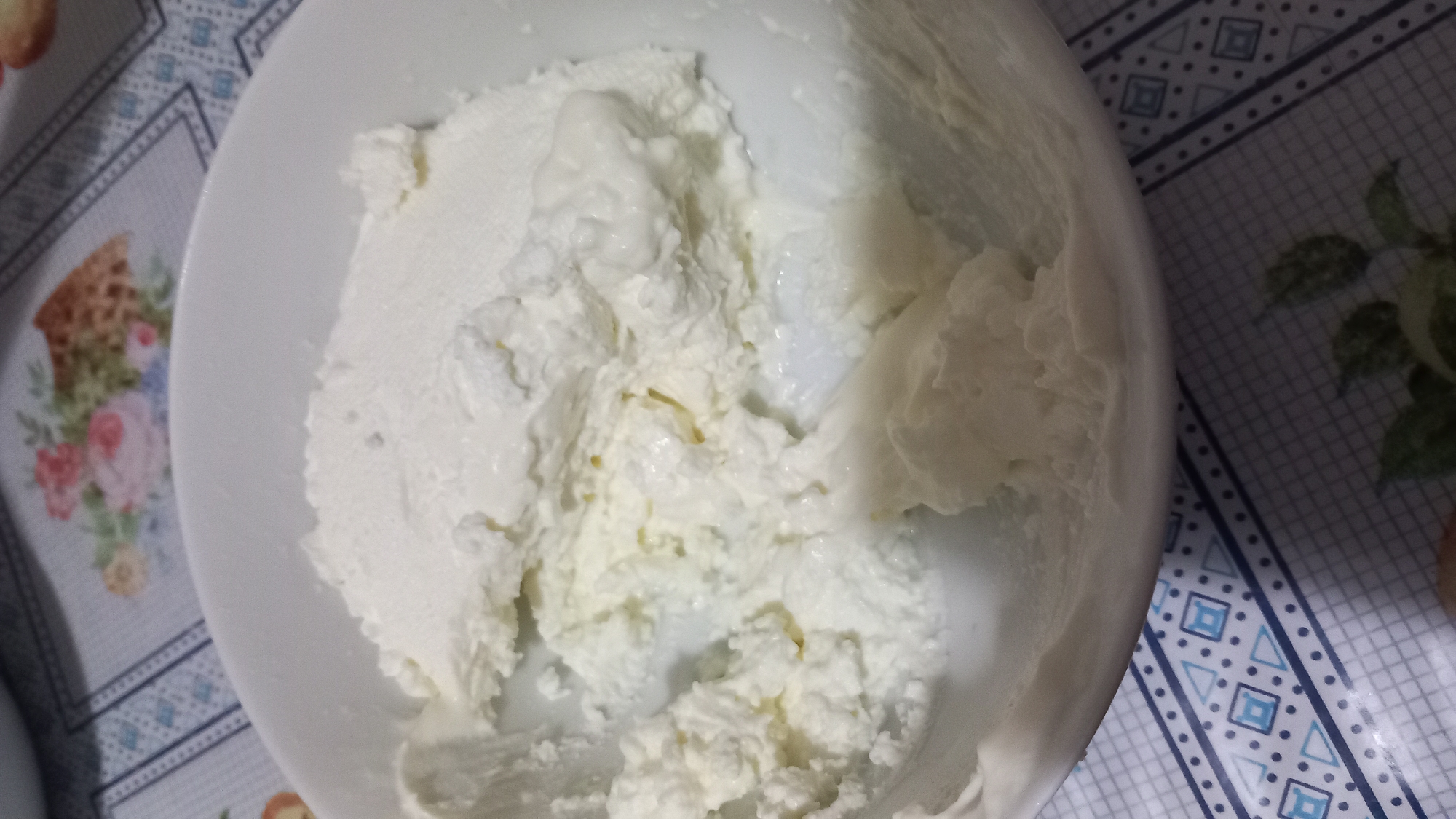 A close-up shot shows a white bowl filled with a creamy, white substance that appears to be ricotta cheese. The substance is textured, with some areas appearing smoother and others more clumpy. The bowl is placed on a table with a patterned tablecloth. The tablecloth has a floral pattern in the upper portion of the image and a geometric pattern in the lower portion. The scene is likely indoors. The location is not specified in the image or crops.
