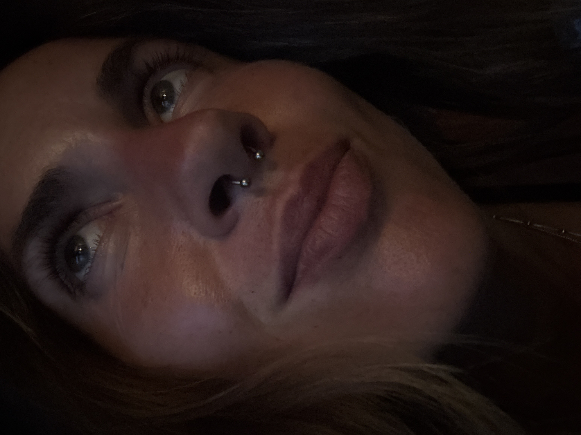 The media file is a close-up, low-angle, portrait-style photograph of a woman's face, taken in a dimly lit environment. The subject's head is tilted, and she is looking upwards and slightly away from the camera. Her hair, which appears to be dark and somewhat long, frames her face and falls into the background.
Notable details include a septum piercing with a horseshoe-shaped barbell. Her eyes are visible, with one slightly more in focus than the other. The lighting casts shadows and highlights on her skin, emphasizing the contours of her face, including her cheekbones, nose, and lips. The overall mood conveyed is intimate and perhaps a bit contemplative, due to the close proximity and the subdued lighting. There is no visible text in the image. The time of day and weather are not discernible, and the location is unknown.