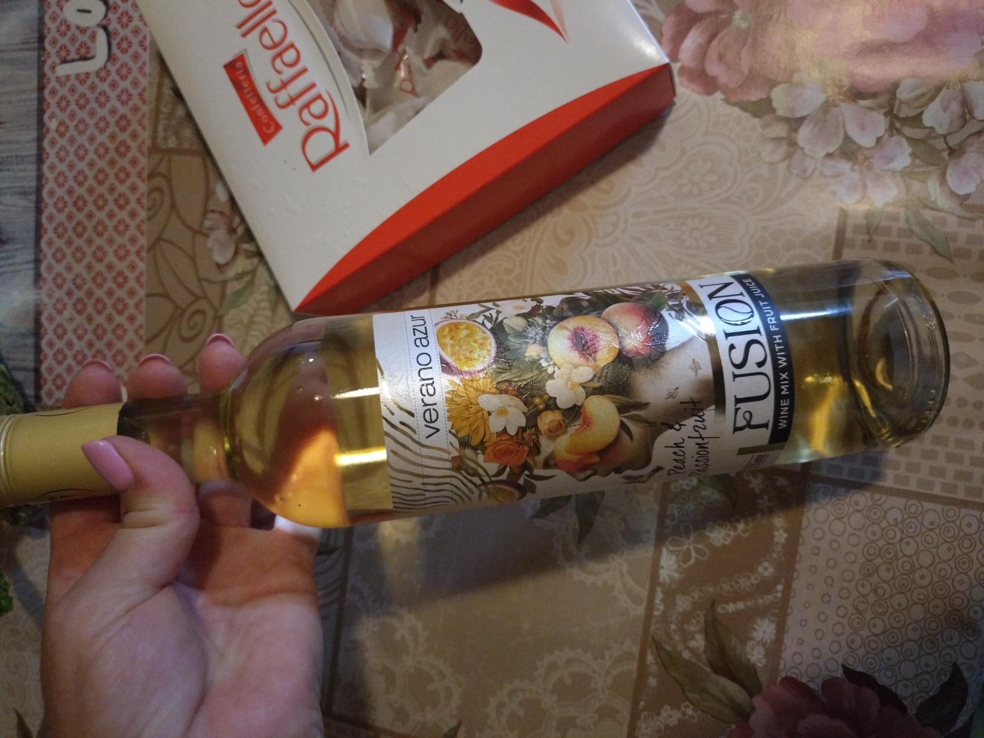 The image shows a close-up of a bottle of "Fusion Wine Mix with Fruit Juice" and a box of "Raffaello" chocolates. A hand with pink fingernails is holding the neck of the wine bottle. The wine bottle has a label with floral and fruit illustrations, and the text "Verano Azur" and "Peach & Passionfruit". The Raffaello box is white with red accents and the brand name is clearly visible. The objects are placed on a patterned tablecloth. The scene is indoors, likely on a table. There is no indication of the city of Stara Zagora, Bulgaria, in the image.