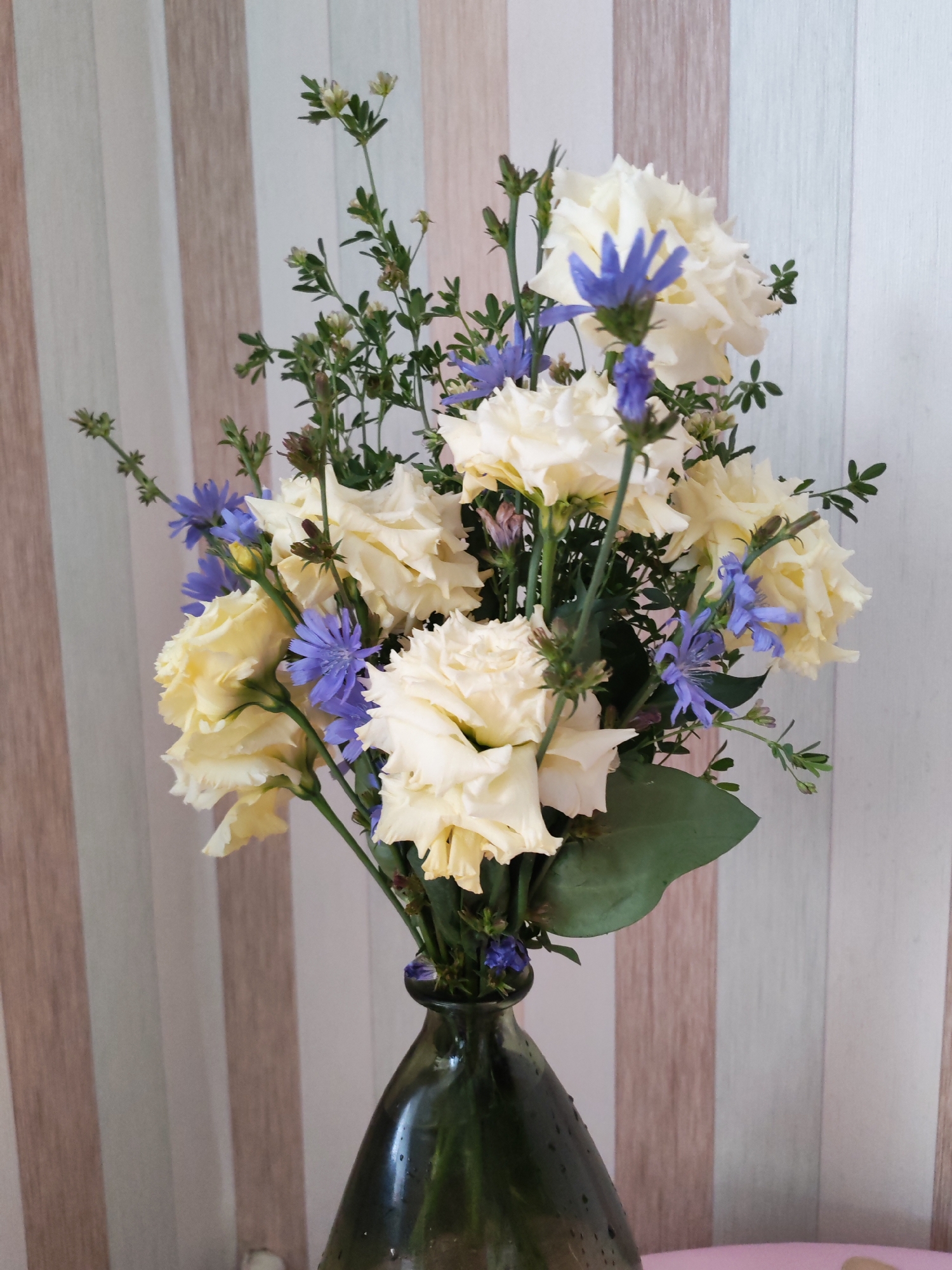 A close-up, vertical shot captures a bouquet of cream-colored flowers with delicate purple accents, artfully arranged in a dark green glass vase. The vase, with a textured, slightly wet exterior, rests on an unseen surface. Behind the bouquet, a wall displays a subtle pattern of vertical stripes in varying shades of beige, white, and a hint of pale pink. The lighting appears soft, casting gentle shadows and highlighting the textures of the flowers and vase. There is no visible indication of the city of Izhevsk, Russia, or any specific location beyond the immediate scene.