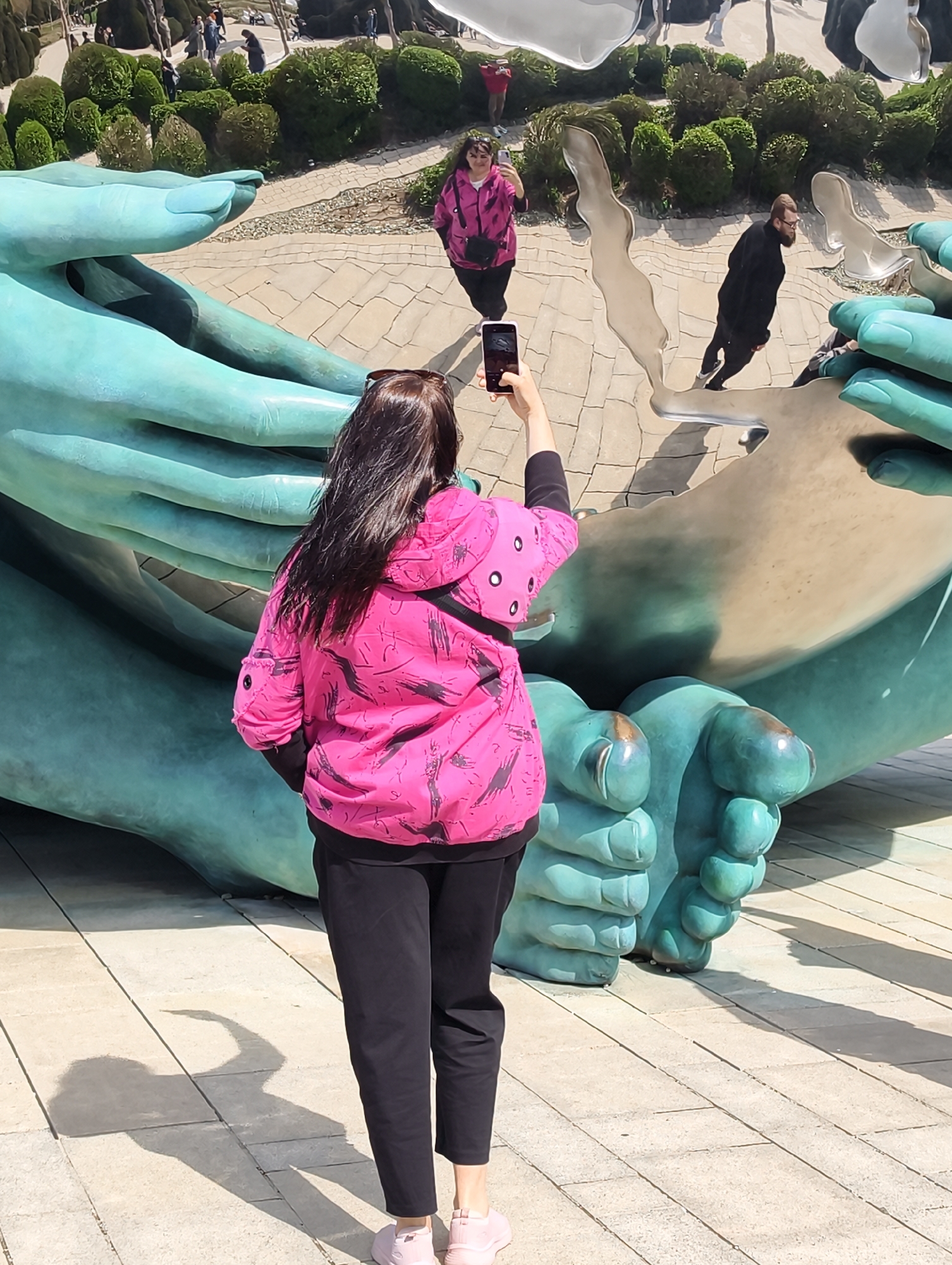 The photo depicts a woman in a pink jacket and black pants taking a photo with her cell phone of a large, teal-colored sculpture of two hands holding a mirrored globe. The mirrored globe reflects the surrounding environment, including a woman in a pink jacket, a man in a black coat, trees, and other people. The ground is paved with light-colored bricks. The photo was taken in Krasnodar, Russia.