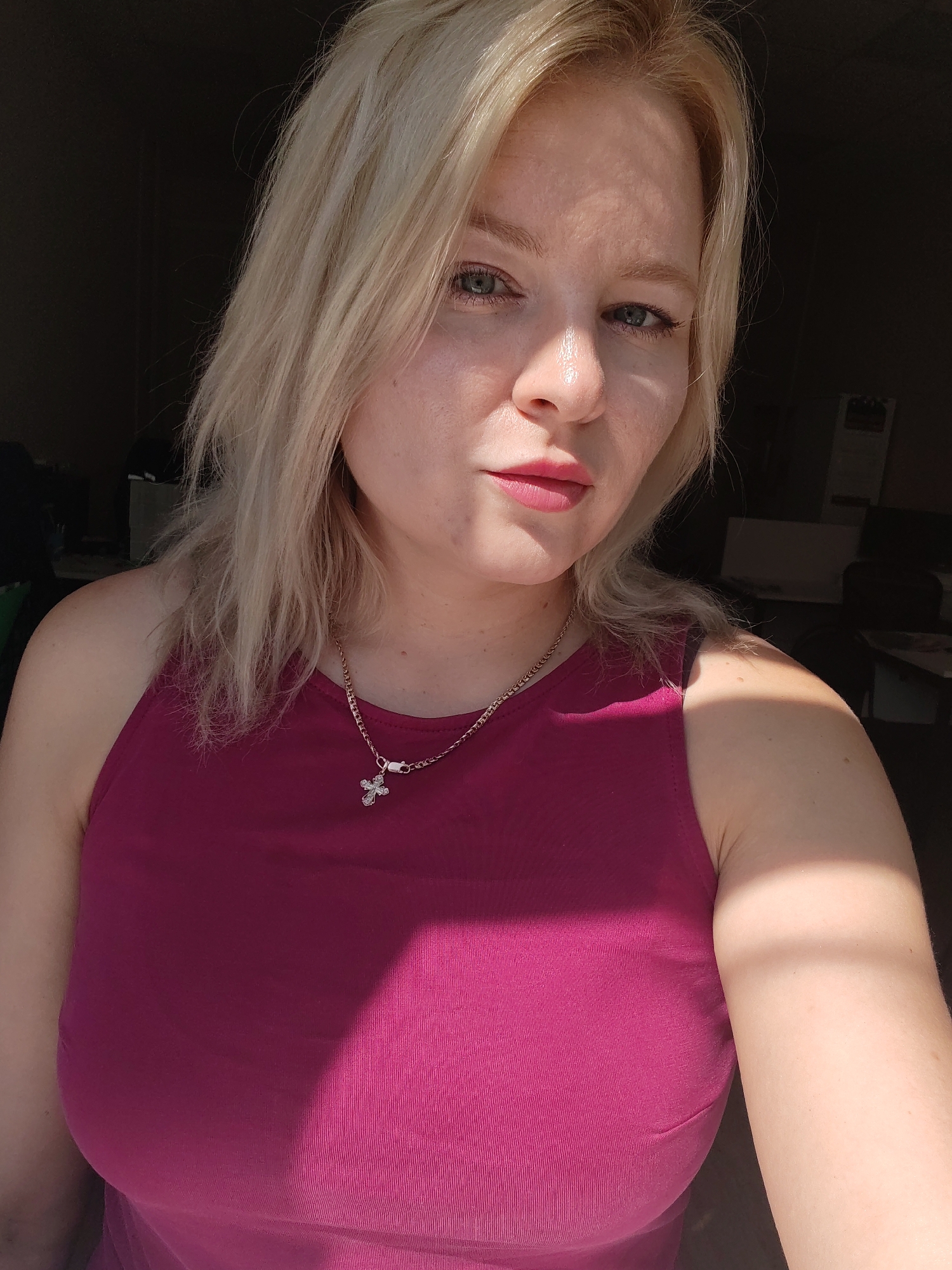 A blonde woman is photographed indoors, looking directly at the camera. She is wearing a magenta tank top and a silver necklace with a cross pendant. Her hair is shoulder-length and styled loosely. The lighting suggests she is near a window, with sunlight casting shadows on her face and chest. The background is dark and out of focus, with hints of furniture and possibly office equipment. The overall impression is a casual, personal photograph. The location in Izhevsk, Russia is not identifiable from the visual information provided in the image.