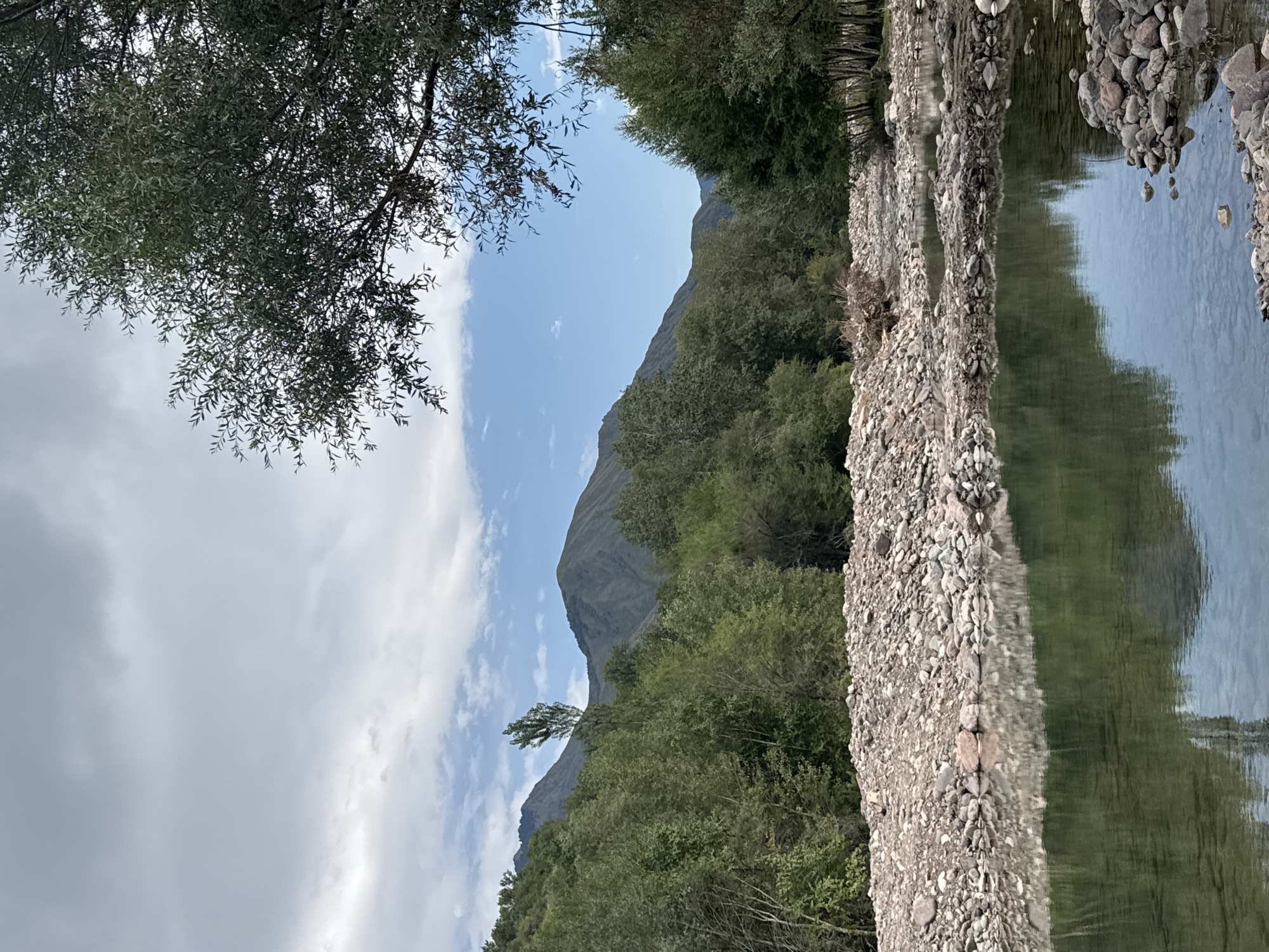 The image shows a natural landscape with a river in the foreground and mountains in the background. The river is shallow and clear, with a rocky bed visible through the water. The banks of the river are lined with stones and pebbles. Beyond the river, there is a dense forest of green trees. In the distance, large mountains rise, their slopes covered in vegetation. The sky is partly cloudy, with patches of blue visible between the clouds. The scene appears to be a tranquil and natural setting. The location is identified as Leskhoz, Kazakhstan.