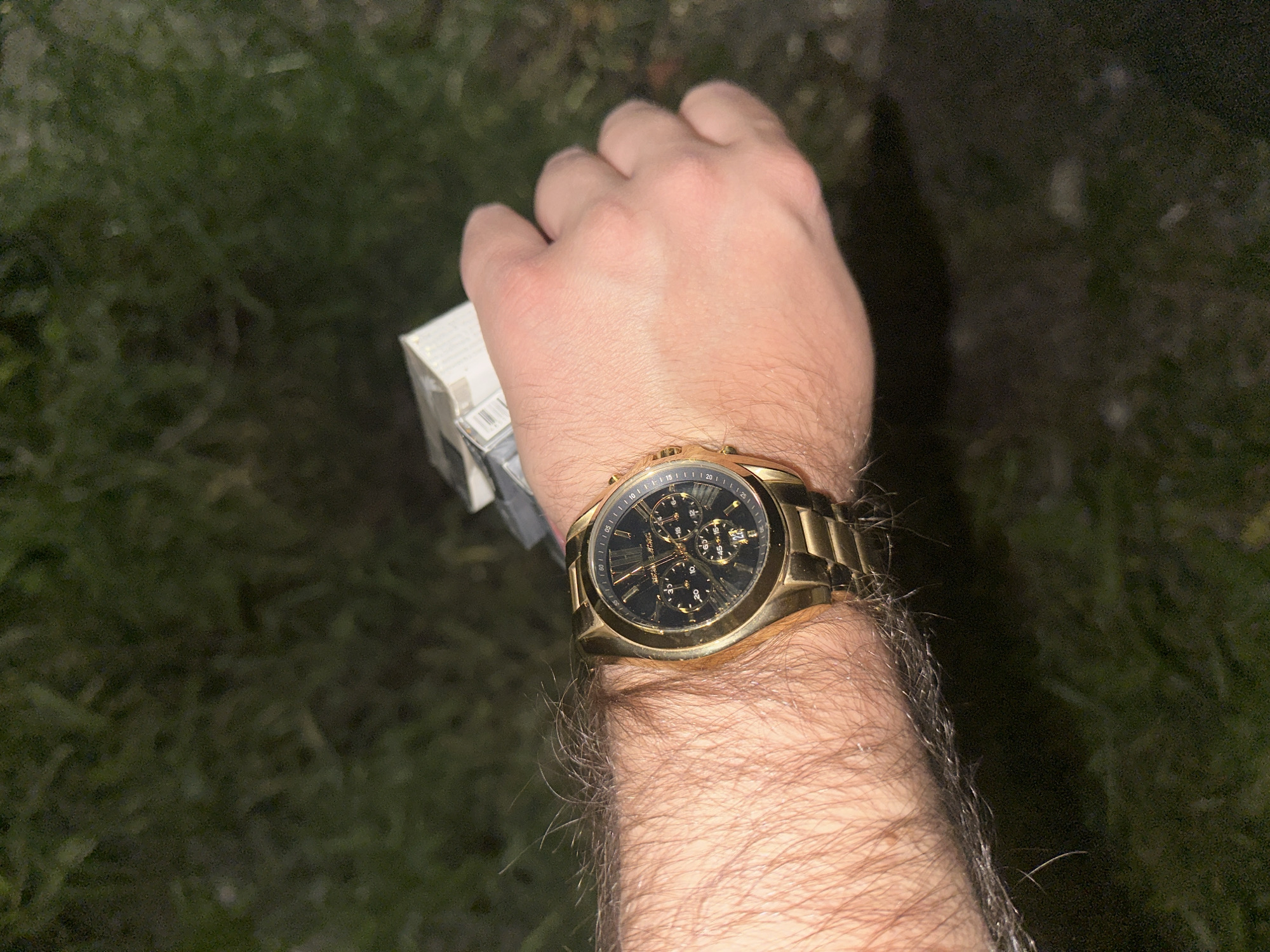 A person's arm and hand are prominently displayed against a dark, indistinct background of green and brown that could be grass or foliage. The individual is wearing a gold-colored wristwatch with a black face and chronograph dials. A white rectangular object, possibly a pack of cigarettes, is held within the hand. The arm is noticeably hairy. The setting appears to be outdoors, possibly at night or in very low light. There is no indication of Yerevan, Armenia in the image.
