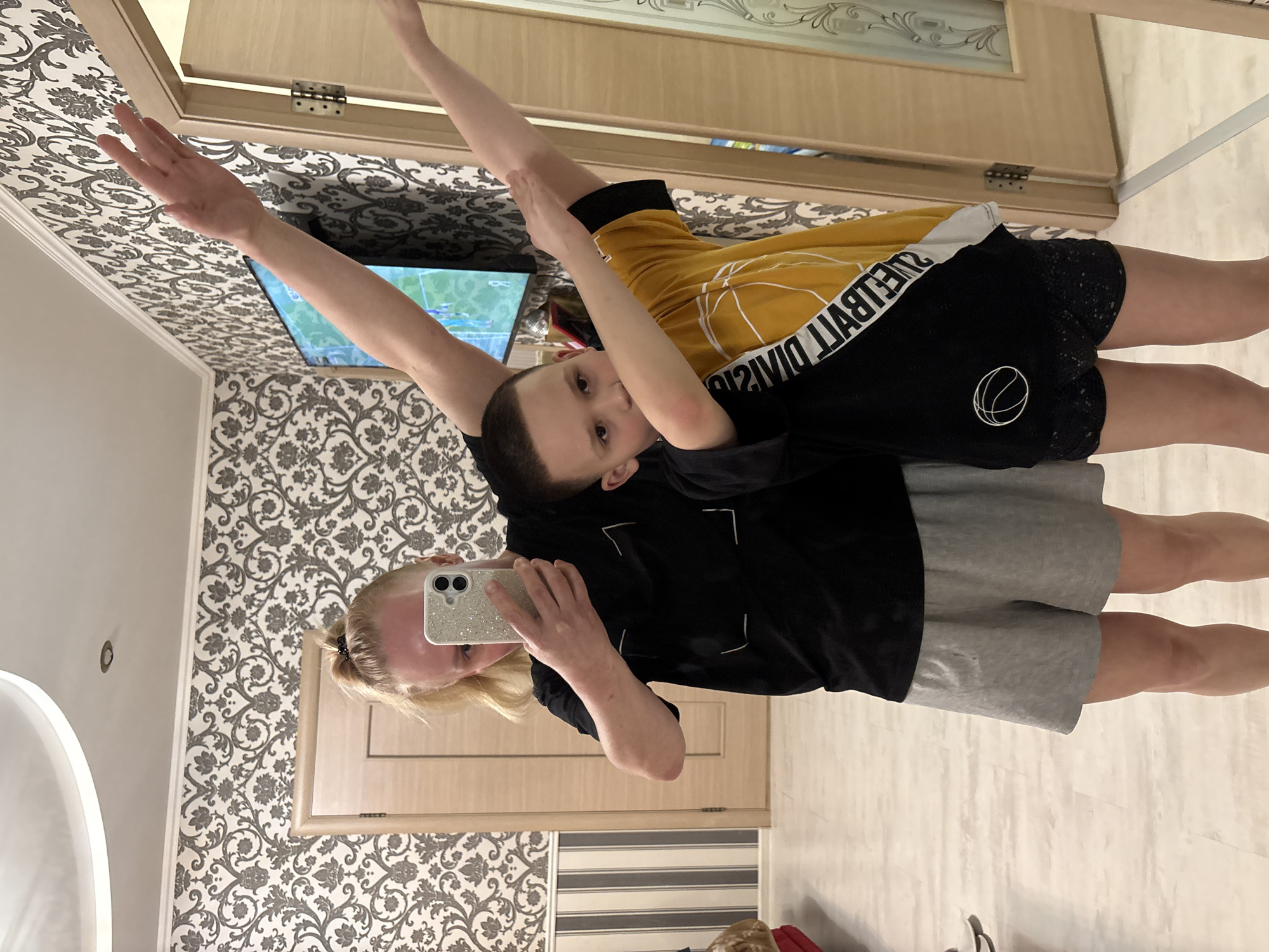 Two individuals are positioned in front of a mirror, seemingly taking a photo. The individual in the foreground is holding a smartphone, possibly capturing the scene. The second individual, a child, stands behind with one arm raised. The child is wearing a black and yellow shirt, and black shorts with a white basketball logo. The first individual is wearing a black shirt, and gray shorts. The location appears to be indoors, based on the wallpaper and flooring visible. A television is partially visible on the wall. The location is described as the city of Chaikovsky, Russia.
