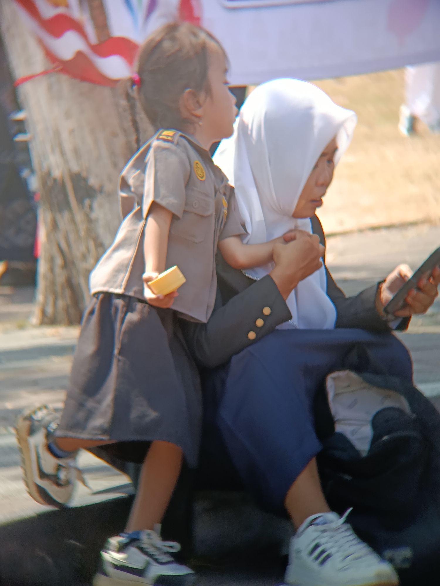 The image captures a portion of an outdoor scene, possibly in Desa Kebon Manis Kecamatan Cilacap Utara, Indonesia, based on the provided context. Two individuals are the primary focus. In the foreground, a young child, dressed in what appears to be a police uniform with a gray shirt and dark skirt, holds a small yellow object in their left hand. The child's right arm is around the neck of the second individual. This second individual, likely an adult woman, is wearing a white hijab and a dark blazer over a blue skirt. She is looking down and holding a mobile phone in her right hand, with her left arm supporting the child. The background is blurred but suggests an outdoor setting with a tree trunk visible on the left and dry, grassy ground. Sunlight illuminates the scene, casting soft shadows. The overall impression is a candid moment of interaction between the two people.
