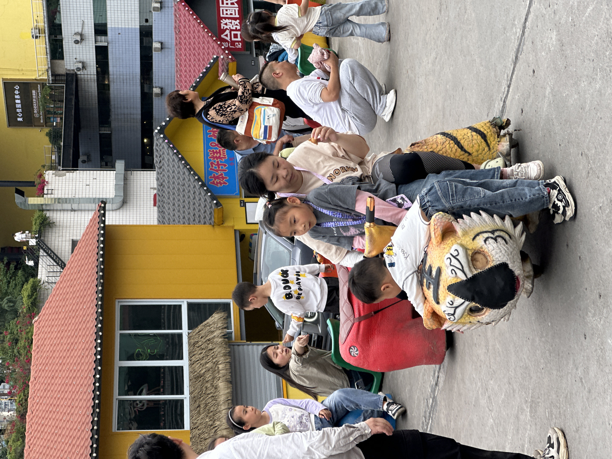 Here's a detailed description of the image:
**Scene:** The image appears to be taken in a public area, possibly a plaza or wide sidewalk in Nan'an District, China. The setting includes vibrant architecture, suggesting a commercial or recreational area. A number of people are gathered, some sitting on what appear to be novelty animal-shaped rides.
**People:**
* There are adults and children present. The adults seem to be in a supervisory or accompanying role.
* The children are mostly interacting with the animal-shaped rides, either sitting on them or near them.
* Some adults are holding or using cell phones, possibly taking photos or videos of the children.
**Objects:**
* The most prominent objects are the animal-shaped rides, particularly those resembling a tiger and other unidentified animals. They appear to be designed for children to sit on and possibly move around.
* The architecture in the background features bright colors like yellow and red, and some have what looks like signage with Chinese characters.
* Other objects include clothing, bags, and accessories worn by the people.
**Actions and Interactions:**
* The children are primarily focused on the animal rides.
* The adults are either looking at the children or using their phones.
* The overall atmosphere seems relaxed and informal, with families enjoying leisure time.
**Environment:**
* The environment is urban.
* The architectural style is unique with vivid colors, which contribute to the cheerful atmosphere.
* The ground is paved, and the weather appears to be overcast.