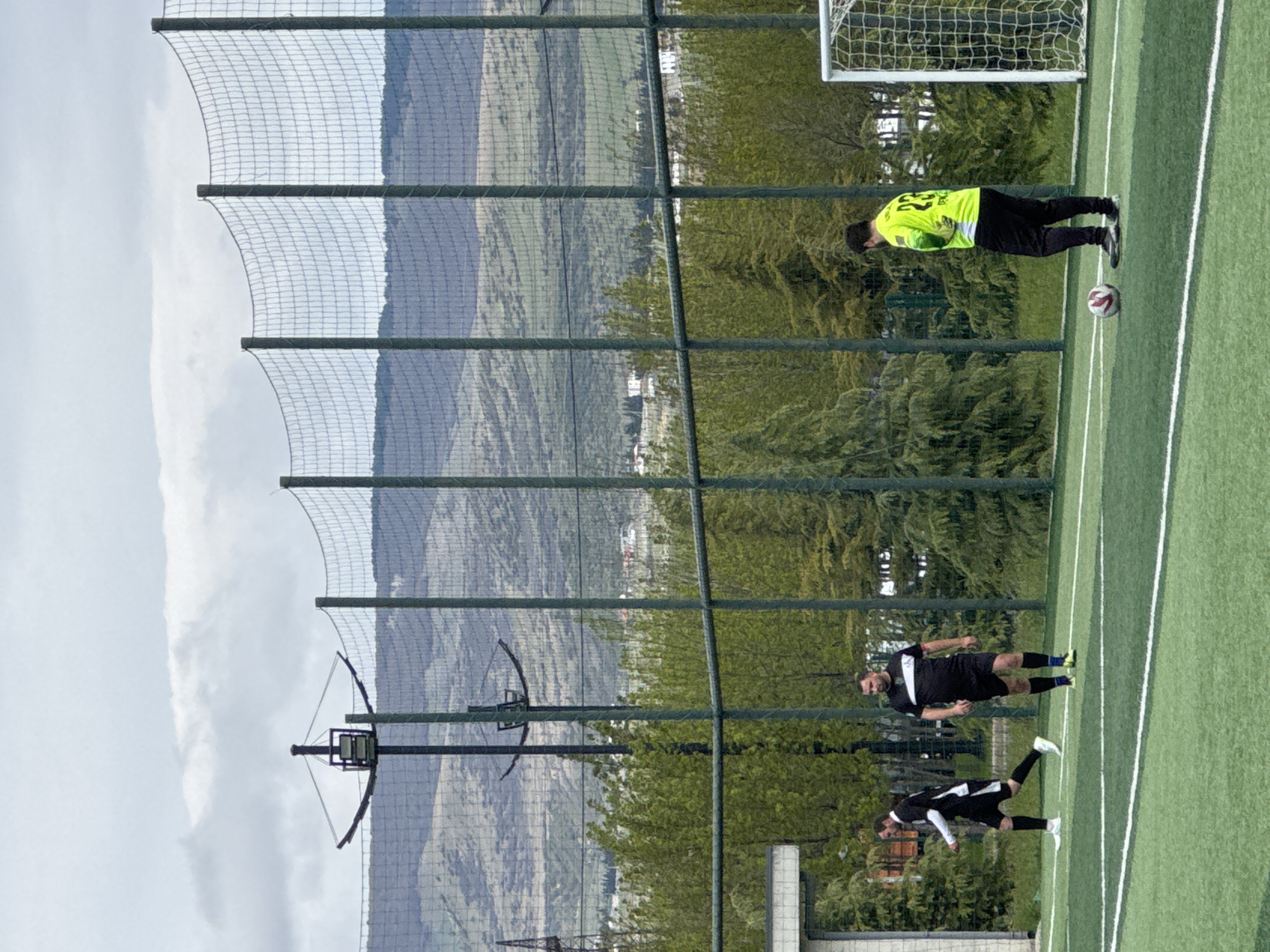 A slightly rotated, eye-level, outdoor shot shows a soccer field in Tbilisi, Georgia.
In the foreground on the right side of the frame, two soccer players are wearing dark jerseys and white socks with dark shoes. One is standing and the other is on the ground. Further up on the right is another soccer player, in a neon yellow shirt, dark pants, and dark shoes, standing near a soccer ball. Behind the neon player is a soccer goal.
A tall, dark metal fence runs the length of the frame on the left. In the background behind the fence, is a wide hillside with trees. Behind the hillside, the sky is overcast.
In the lower left, is a smaller metal fence with a round antenna mounted on it.