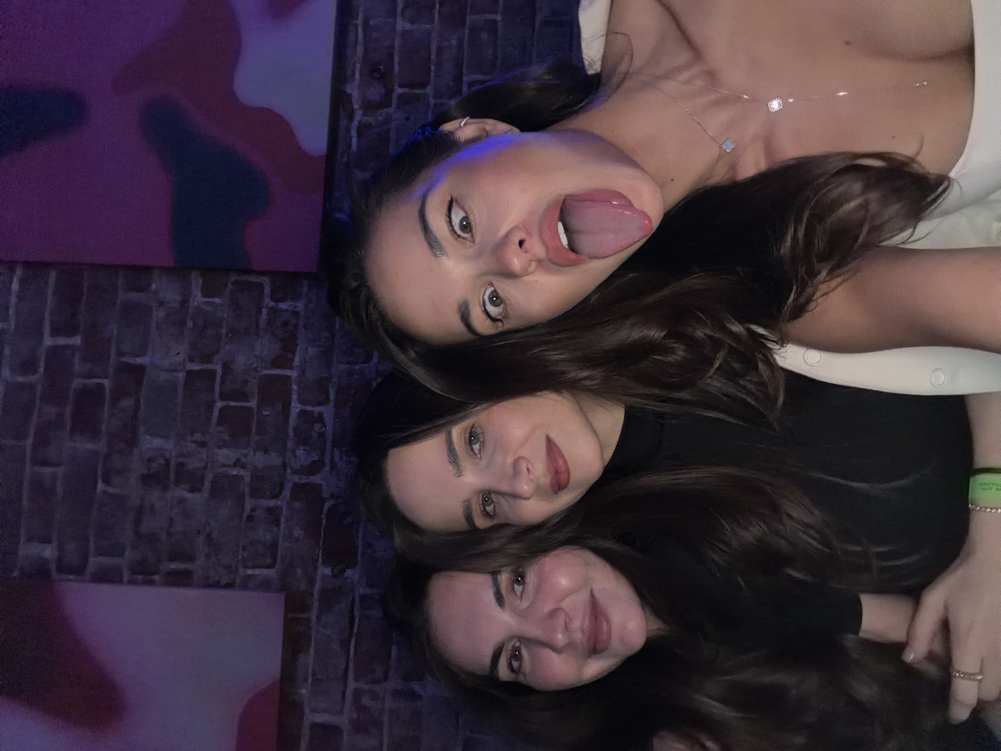 The image, taken in Krasnoyarsk, Russia, features three young women captured in a close-up, likely lying down or leaning very close to each other. Their faces are prominent in the foreground, with their dark hair flowing around them.
The woman positioned at the top (when the image is correctly oriented) has dark hair and is wearing a light-colored top. She is looking directly at the camera with wide eyes and playfully sticking out her tongue. A delicate silver necklace with two small charms, one square and one resembling a four-leaf clover, is visible around her neck. She also wears an earring on her left ear.
Below her, the second woman, also with dark hair, is wearing a dark top. She has a gentle smile on her face. A green wristband is visible on her left wrist, though the text on it is not clearly legible.
At the bottom of the frame, the third woman, with dark hair, wears a dark top and smiles warmly at the camera. Several rings are visible on the fingers of her right hand.
The background consists of a dark brick wall. To the left, an abstract piece of art featuring soft-edged shapes in shades of pink, purple, and dark blue is visible. The lighting is dim, suggesting an indoor evening or nighttime setting, possibly in a bar, lounge, or a similar relaxed venue. The overall mood is casual and friendly, capturing a moment of shared enjoyment among friends.