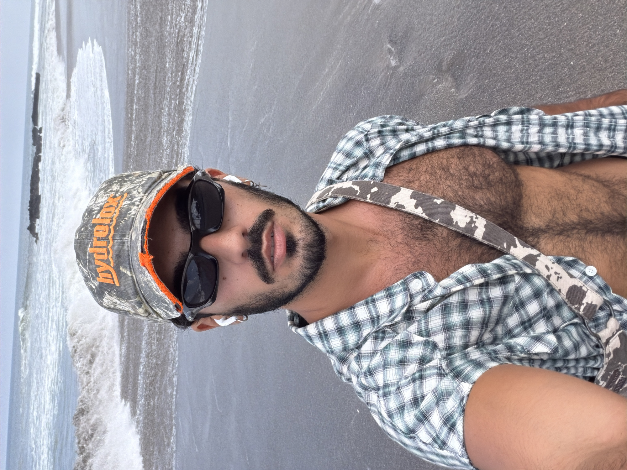 A man stands on a dark sand beach in North Kuta, Indonesia, facing the camera. He wears a camouflage baseball cap with orange trim, dark sunglasses, and a plaid short-sleeved shirt unbuttoned, revealing his chest. A strap with a cow-print pattern crosses his chest. The ocean with white waves is visible in the background.