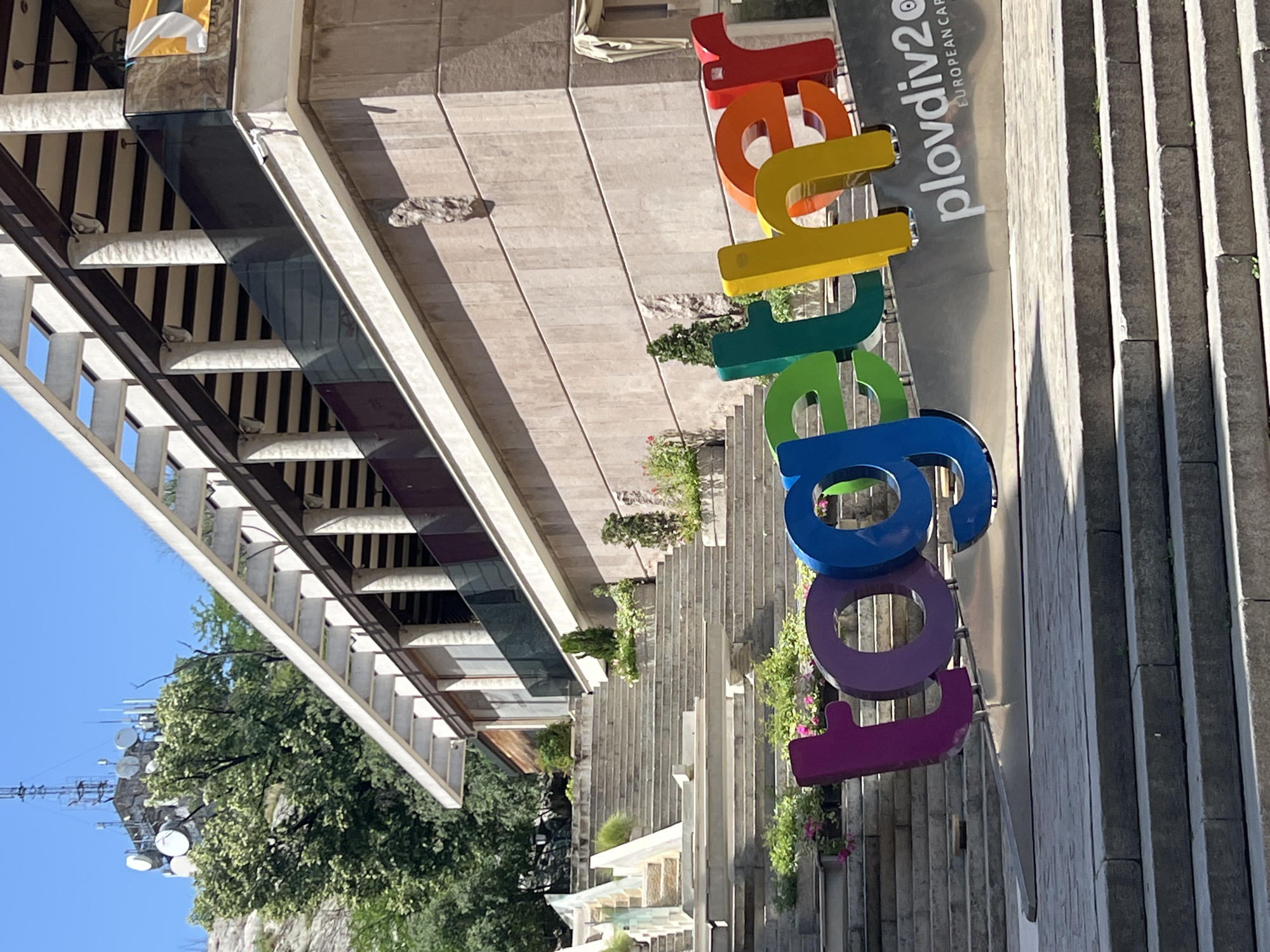 The image captures a view in Plovdiv, Bulgaria, featuring a large, colorful sign that spells out "Plovdiv2019 European Capital of Culture". The letters are arranged vertically with red at the top, followed by orange, yellow, green, blue, and purple. The sign is positioned adjacent to a set of stone stairs leading up to a building with a modern architectural design. The building has multiple levels with glass-covered balconies. Vegetation, including trees, bushes, and flowers, adorns the area around the stairs and the building. In the background, a communication tower can be seen against a clear blue sky. The image is taken from a ground-level perspective.