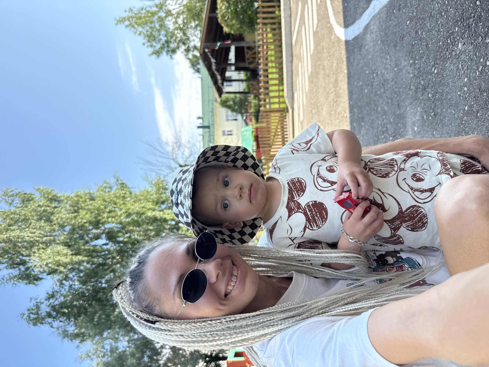 In Rusanovo, Russia, a woman with long blonde braided hair and dark sunglasses is taking a selfie with a young child. The woman is wearing a white t-shirt. The child is wearing a checkered bucket hat and a light-colored t-shirt with a repeating brown Mickey Mouse pattern. The child is holding a small red toy car. They are standing outdoors in what appears to be a playground or park area with a paved surface marked with white lines, suggesting a crosswalk. In the background, there is a wooden gazebo, a low wooden fence, green trees, and buildings. The sky is blue with some scattered clouds.