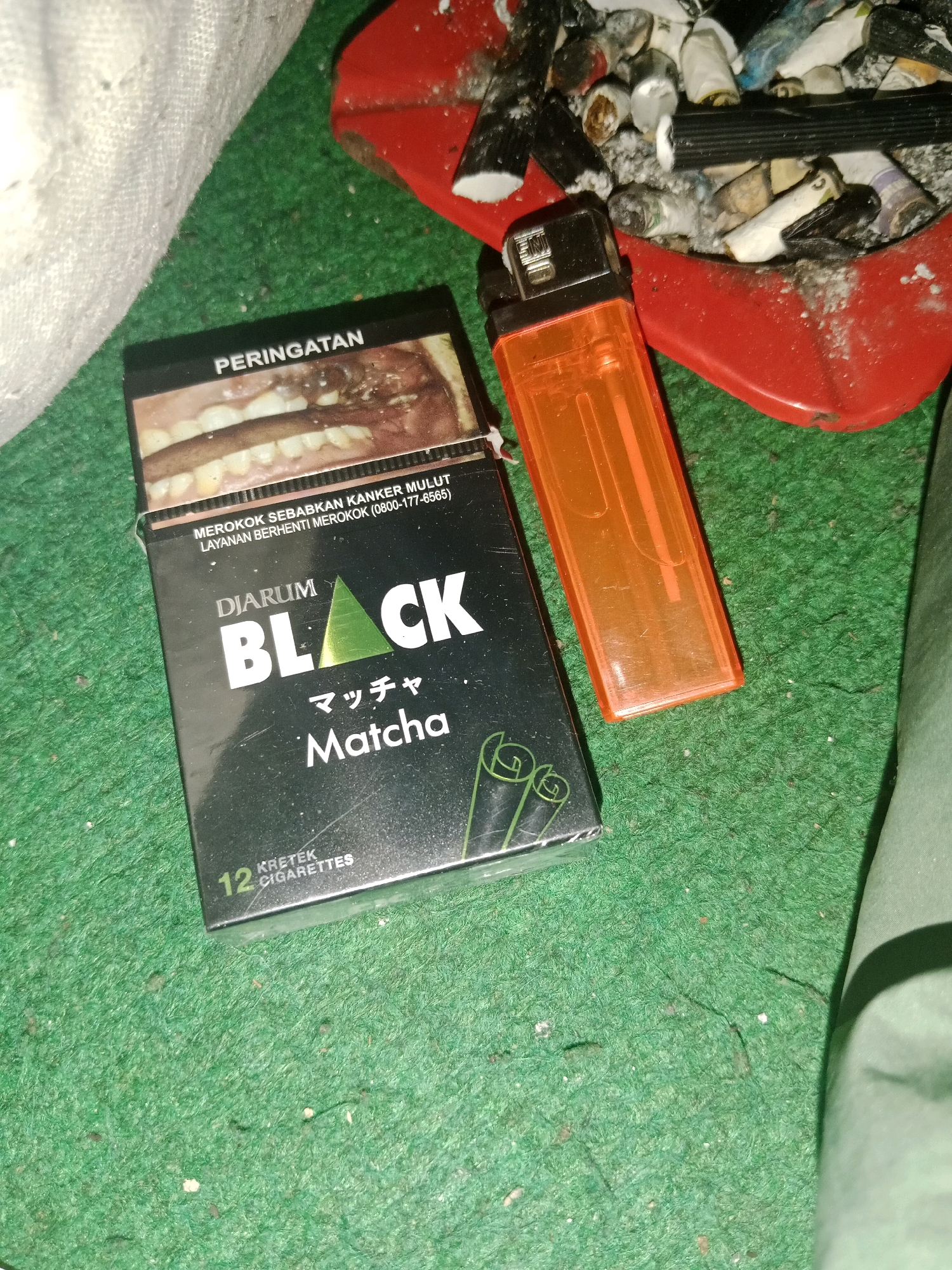 The image shows a close-up of a pack of Djarum Black Matcha kretek cigarettes and an orange lighter on a green, carpeted surface. A red ashtray filled with cigarette butts and debris is visible in the background, to the upper right of the cigarettes. The lighting is dim. The scene appears to be indoors, likely a personal space. There is no indication of the location within Ringinrejo, Indonesia.