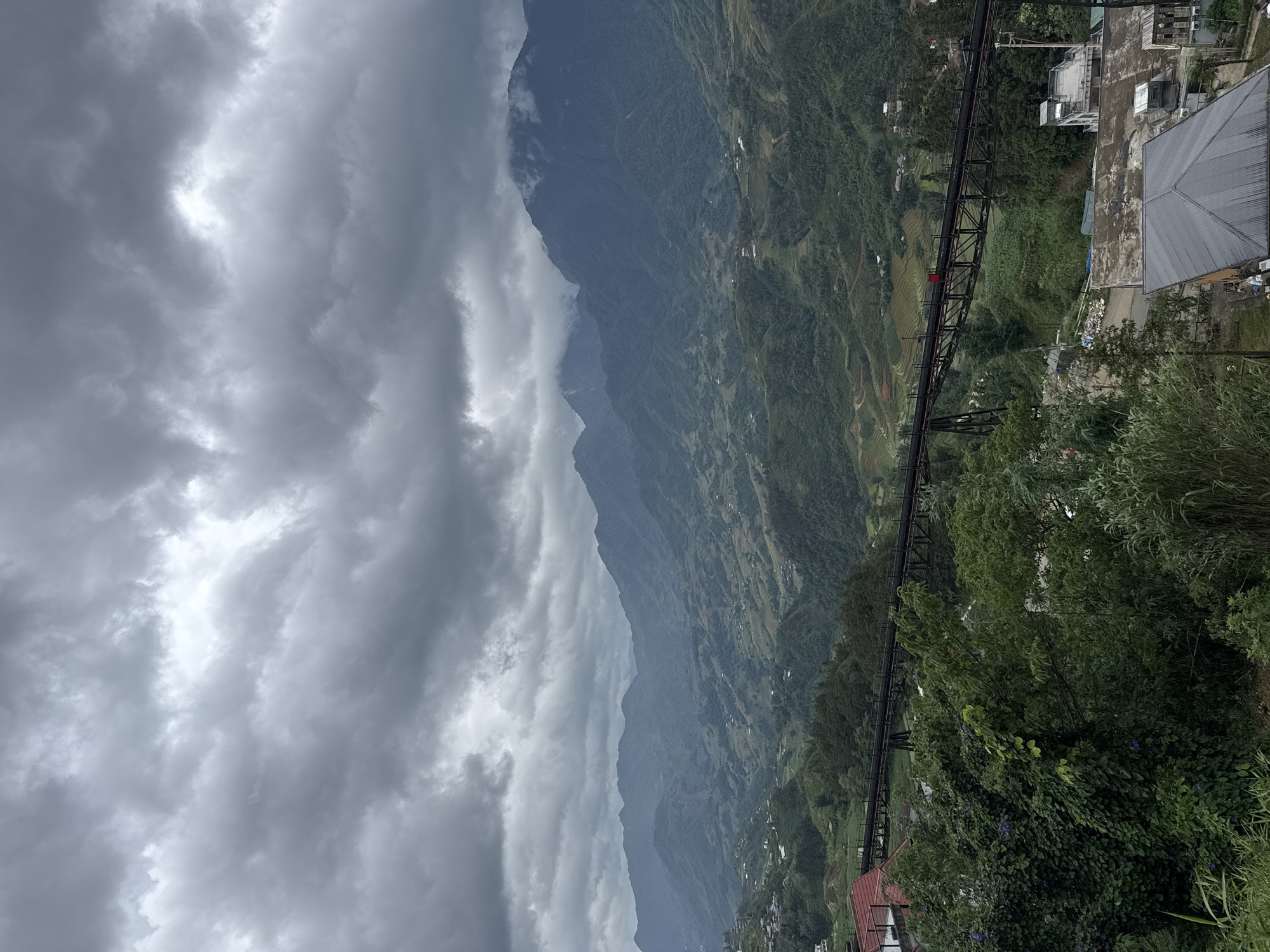 The image captures a landscape from Sa Pa Ward, Vietnam, under a cloudy sky. A mountain range stretches across the horizon, partially obscured by mist. Lush greenery covers the mountain slopes, dotted with what appear to be small settlements or agricultural terraces.
In the foreground, a metal structure, possibly a bridge or elevated walkway, traverses the landscape. Adjacent to this structure, are buildings with rooftops, suggesting a built-up area or town. The lower right corner of the image is dominated by dense foliage, further emphasizing the area's natural environment.