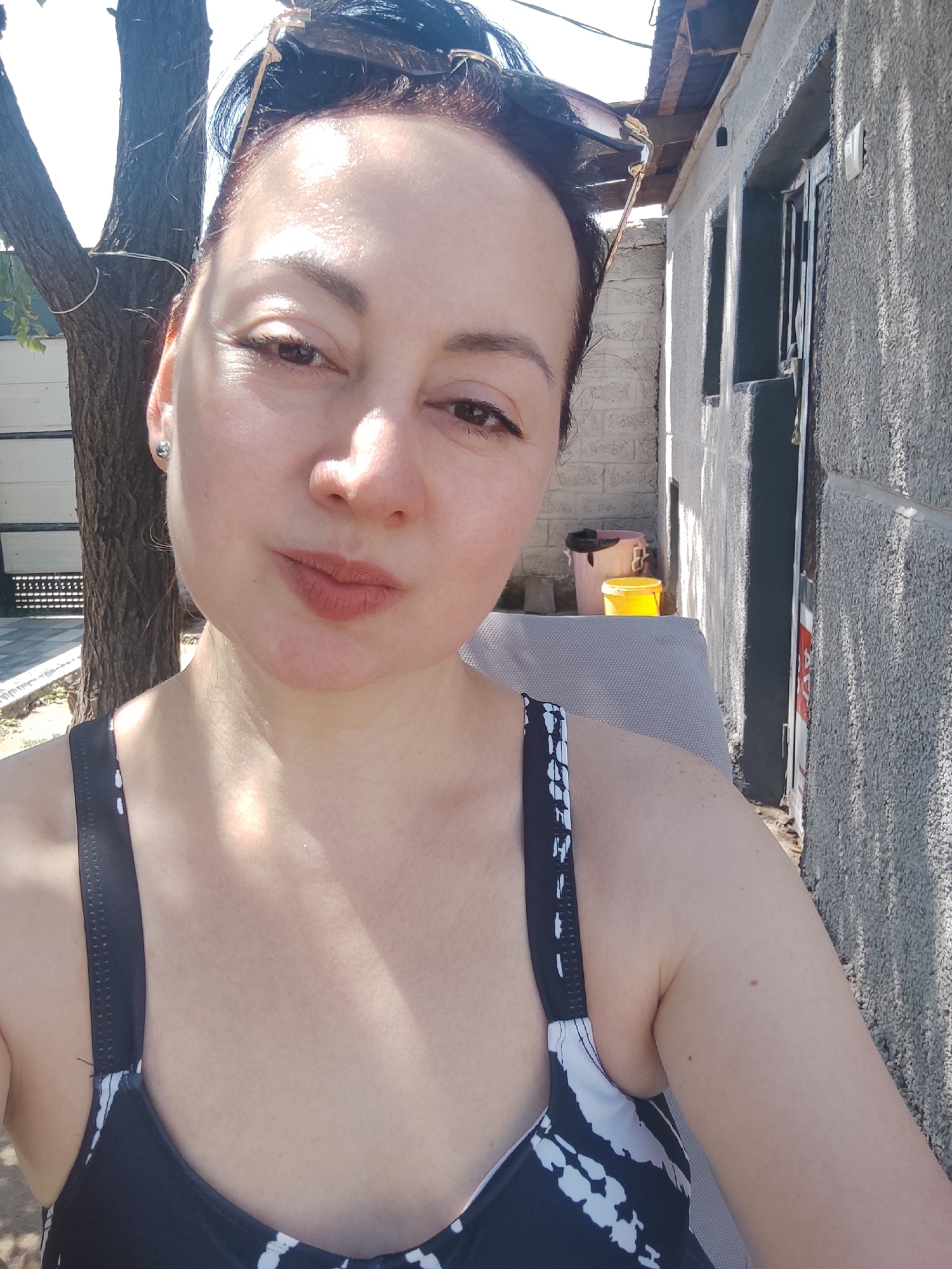 The image is a selfie of a woman in Eshmataul, Uzbekistan, taken outdoors in daylight. She is wearing a black and white patterned swimsuit. The background features a tree, a textured gray wall, and a building with windows and a door. There are two yellow buckets visible near the building. The woman has dark hair and is looking towards the camera with a neutral expression. The lighting suggests it is a sunny day, casting shadows on the wall.