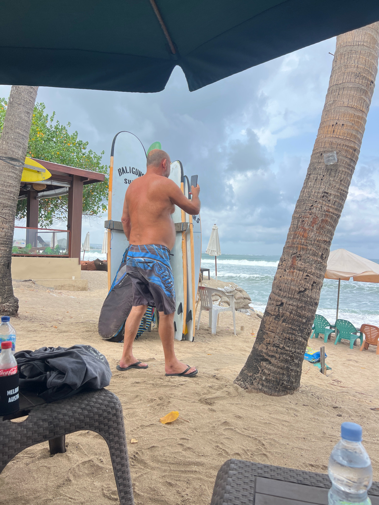 A shirtless man in blue and black swim trunks stands on a sandy beach in Kuta, Indonesia, holding a mobile phone to take a picture. He is positioned in front of several surfboards leaning against a wall, with the ocean and waves visible in the background. Palm trees and beach umbrellas are also present. In the foreground, a table with a black fabric bag and two bottles of water is visible. The sky is overcast.