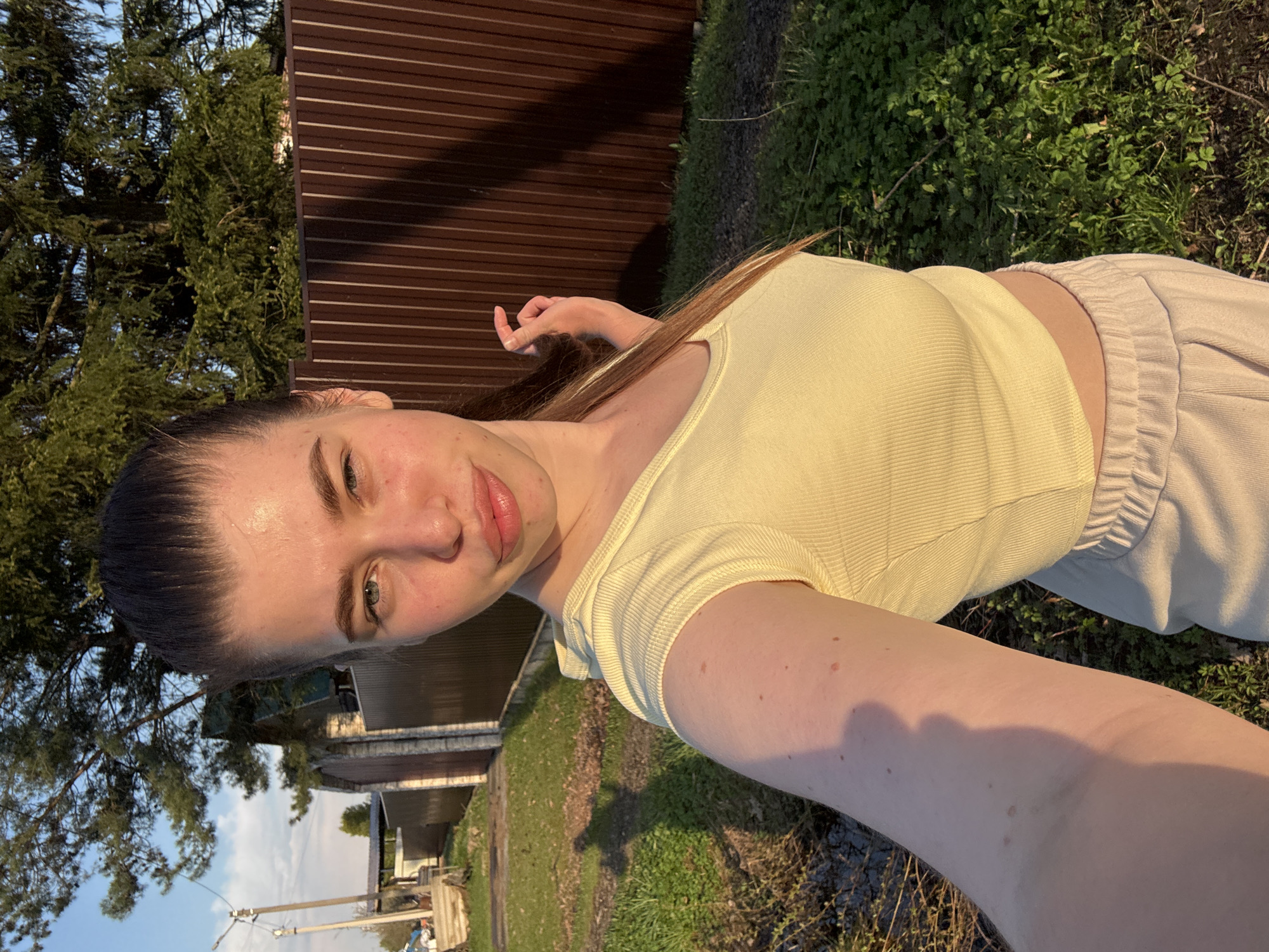 A woman is taking a selfie outside in Шевлягино, Russia. She is wearing a yellow t-shirt and light-colored pants. A brown fence and trees are in the background. Buildings and power poles are visible in the distance, partially obscured by trees. There is grass and vegetation on the ground.