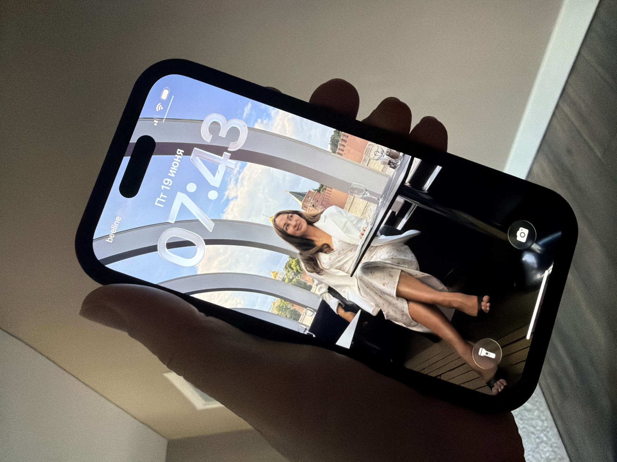 An iPhone is held by a hand, displaying a lock screen with a picture of a woman sitting at a table. The lock screen shows the time as 7:43 and the date as June 19th. The background of the image is a view of a cityscape with red brick buildings and a dome, likely the Kremlin in Moscow, Russia. The phone is positioned in front of a wall and wooden floor.