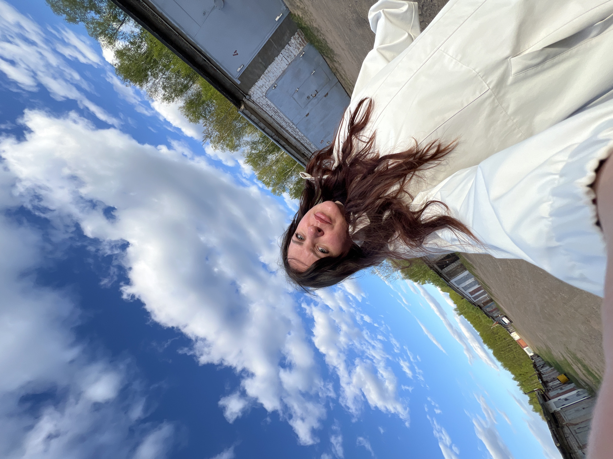 The image shows a person with brown hair, likely female, wearing a white coat. The person is taking a selfie in Noviy, Russia. The background includes a blue sky with scattered white clouds, some trees, and a row of garage doors. The ground appears to be dirt or pavement.