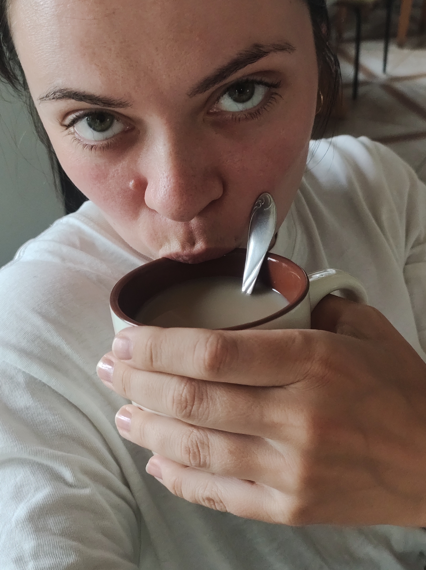 A close-up, high-angle shot features a person, likely a woman, looking directly into the camera while drinking from a mug. She has dark hair, prominent dark eyebrows, and striking green or hazel eyes. Her face shows visible skin texture, including pores, and a slight redness around her nose and cheeks, with a small, raised blemish or mole on her nose.
She holds a cream-colored ceramic mug, which has a brown rim and interior, with both hands as her lips touch the beverage inside. A silver spoon, featuring a decorative pattern on its handle, rests within the light brown or beige liquid, suggesting coffee or tea with milk. Her fingernails are manicured with a clear or very light pink polish. She is wearing a white t-shirt, visible around her shoulders and chest, which has some natural wrinkles.
The background is softly blurred but indicates an indoor setting. Hints of furniture, possibly a table and chair, and a patterned floor are vaguely discernible to the right, suggesting a home or casual interior. The lighting is soft and even, creating a serene mood. The subject's direct gaze and the intimate act of drinking contribute to a sense of a quiet, personal moment. There is no visible text in the image.