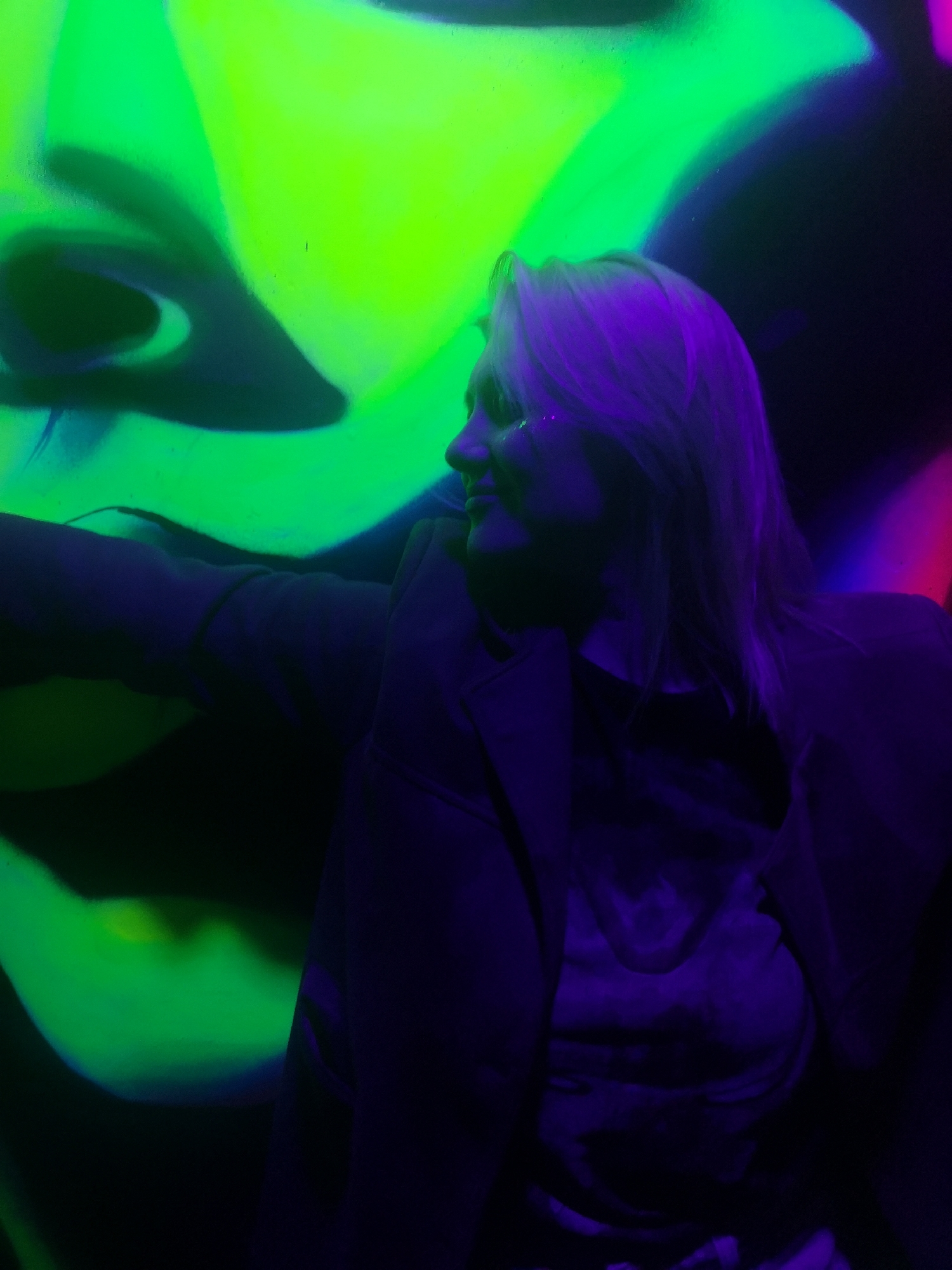A woman with blonde hair is standing in front of a colorful neon mural. The mural appears to depict a stylized face with prominent green and black sections. The woman is wearing a dark jacket over a shirt, and her face is illuminated with a purple glow. It is assumed the photo was taken in Chaikovsky, Russia. There are no other objects, people, or discernible features mentioned in the description.