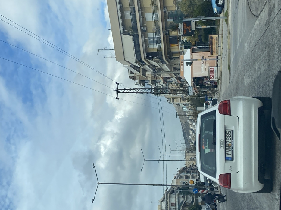 The image presents a daytime street scene in an urban or semi-urban environment, identified as Nikea, Greece. The perspective is from inside a vehicle, looking forward along a road lined with buildings.
The sky is mostly cloudy, with glimpses of blue peeking through. Overhead, numerous power lines traverse the scene, supported by a tall, metal electricity pylon positioned on the left side of the street. Several streetlights are also visible.
Along the right side of the street, multi-story apartment buildings dominate, characterized by balconies, awnings, and air conditioning units. At street level, various establishments are present, including a white kiosk displaying "SMILE KIOSK" and "TSOURAS" (ΤΣΟΥΡΑΣ). Adjacent to it, a traffic light and a bus stop sign with unreadable orange text are seen. Further down, signs for "BARBER SHOP" and "EΛΑΙΟΧΡΩΜΑΤΙΣΜΟΙ" (painting/paint shop) are discernible, along with a Shell station logo.
In the foreground, the rear of a white Audi A3 is prominent. Its Greek license plate reads "YNN-7685," with smaller text "Ν. Κουτλιά" (N. Koutlia) underneath. Other vehicles, including a blue car and a motorcycle, are visible further along the road, suggesting ongoing traffic.
In the background, a range of hills or mountains, featuring some sparse vegetation, stretches across the horizon under the cloudy sky, framing the town. The scene depicts a typical, active day in the area.