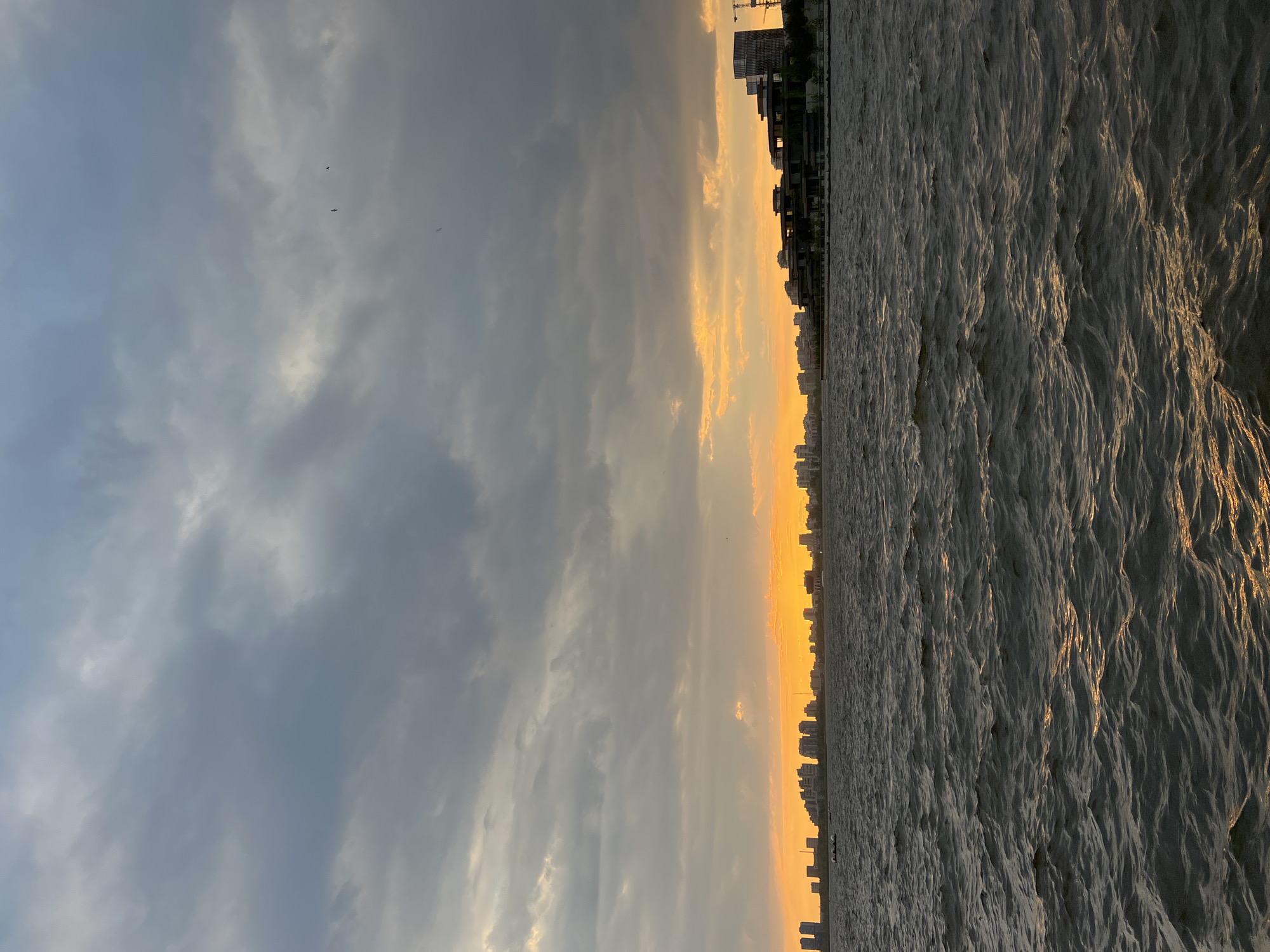 The image depicts a cityscape at sunset. On the right, there's a body of water with a rippled surface. Adjacent to the water is a line of buildings, silhouetted against the setting sun. The sky is filled with clouds, ranging from light gray to dark gray, with hints of orange and yellow where the sun is setting. There are also some birds in the sky. The scene appears to be from Kazan, Russia.