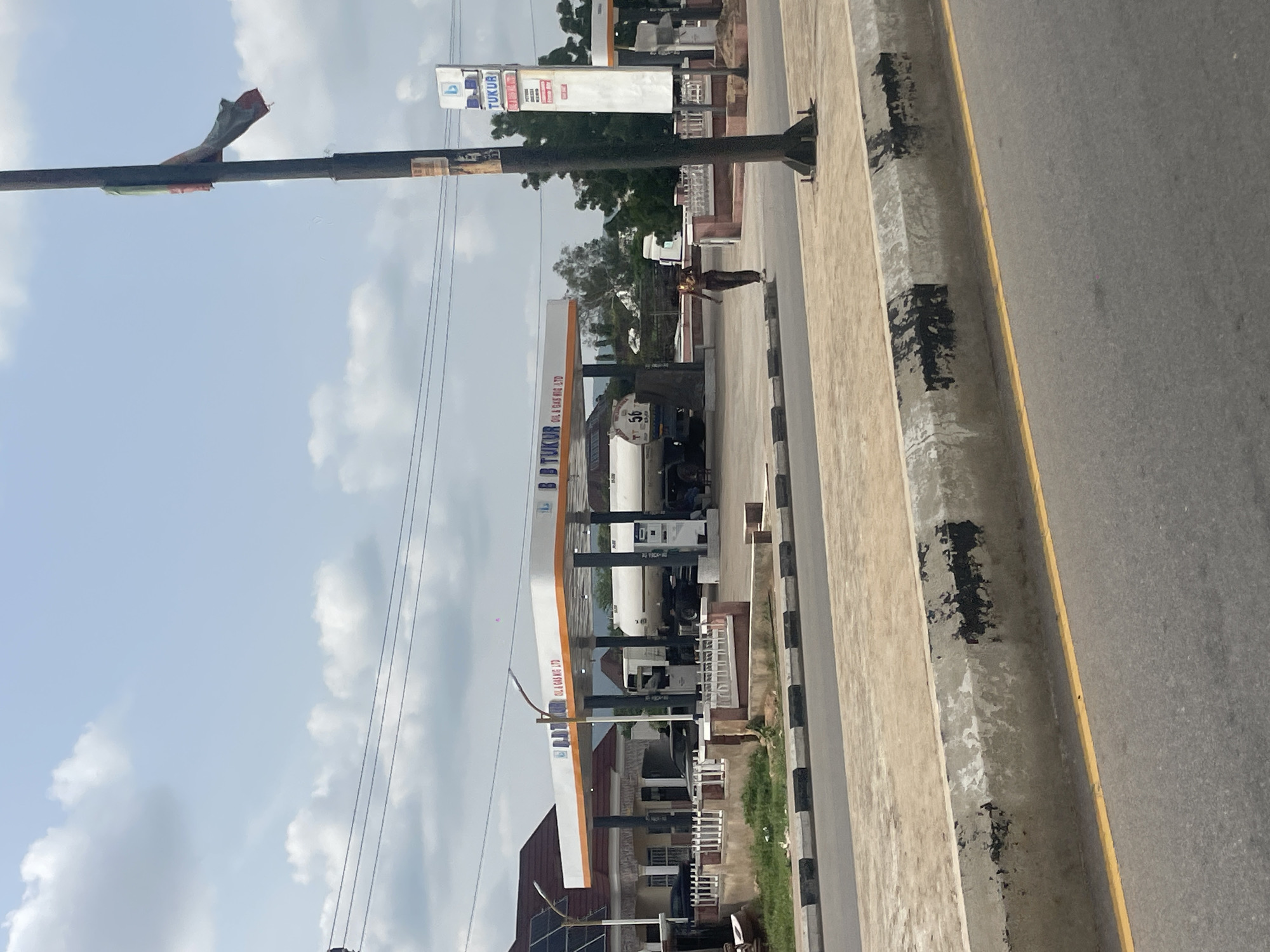 The image depicts a roadside view in Jalingo, Nigeria, featuring a filling station and its surroundings. The prominent structure is a gas station with a white canopy supported by pillars, advertising "BB TUKUR OIL & GAS NIG. LTD." A large tanker truck, labeled with the number "56", is parked at one of the pumps, suggesting active service. Adjacent to the gas station, a building with a tiled roof and covered porch is visible, with cars parked in front. A person, dressed in dark clothing, is walking on the sidewalk in front of the gas station. A tall pole with a tattered flag is positioned on the right side of the image, and a blue and white signage for "TUKUR" is also present. The foreground shows a paved road with a yellow line and a concrete curb, indicating a traffic area. The sky is partly cloudy, with a light blue hue. The overall scene captures a typical commercial area on a street in Jalingo.
