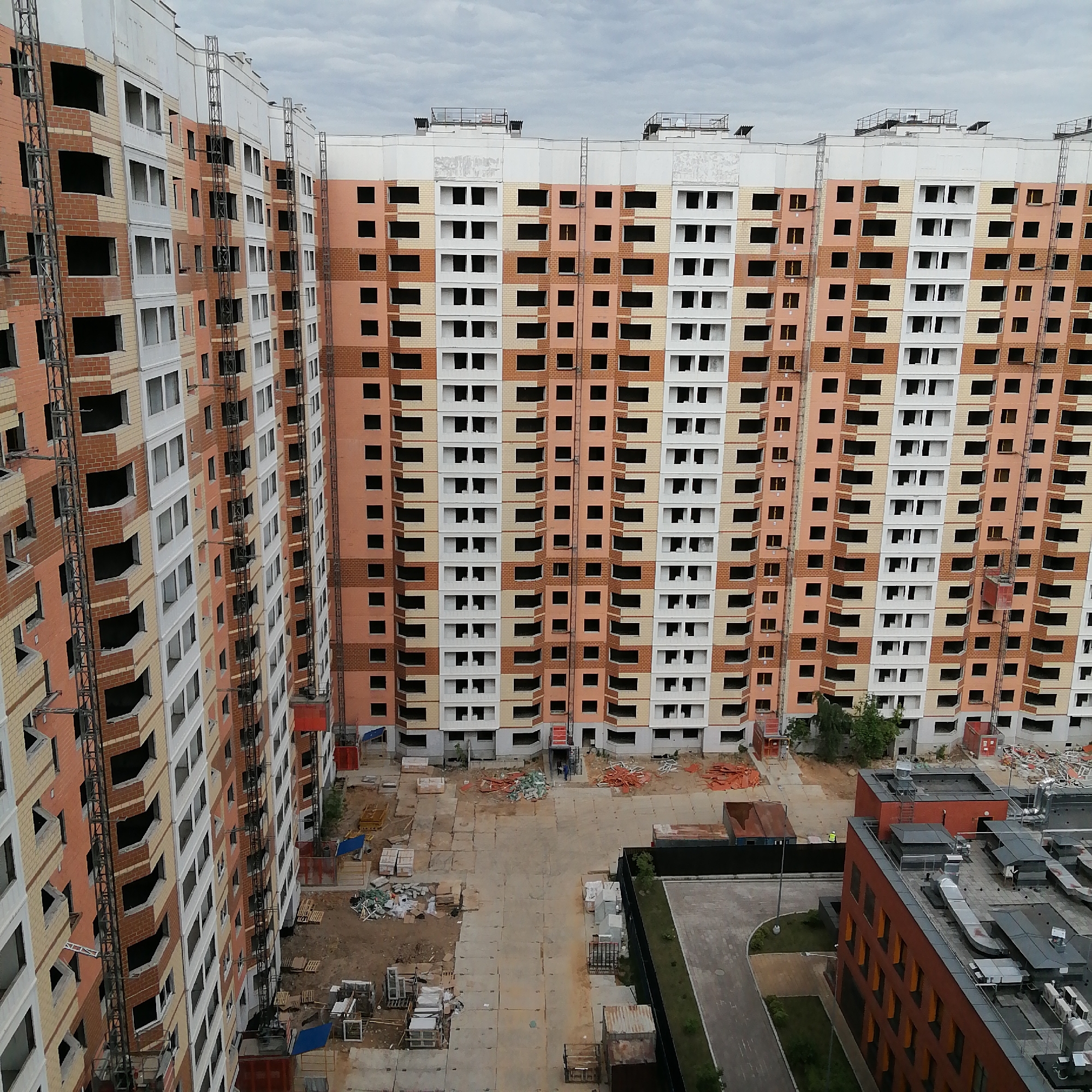 An aerial or high-angle view captures a large construction site featuring multiple high-rise residential buildings. The dominant structures are two long, curving buildings, each with numerous floors, under active construction. Their facades display an alternating pattern of light orange/tan brickwork and white concrete sections, with many window openings still lacking glass.
Vertical metal scaffoldings or hoist systems are affixed to the exterior of the buildings, and several red material lifts are visible ascending or descending these structures. The ground level between the buildings is an expansive, unpaved construction yard, covered with dirt, concrete slabs, and piles of building materials such as pallets, bricks, and various debris.
In the bottom right portion of the image, a completed or nearly completed building with a dark roof, featuring multiple ventilation units and industrial components, is partially visible. Adjacent to this building are a paved road and some landscaped areas with greenery. A single individual wearing a yellow safety vest is discernible near the lower building. The scene unfolds under an overcast sky, indicating daytime with cloudy weather. No legible text is visible in the image.