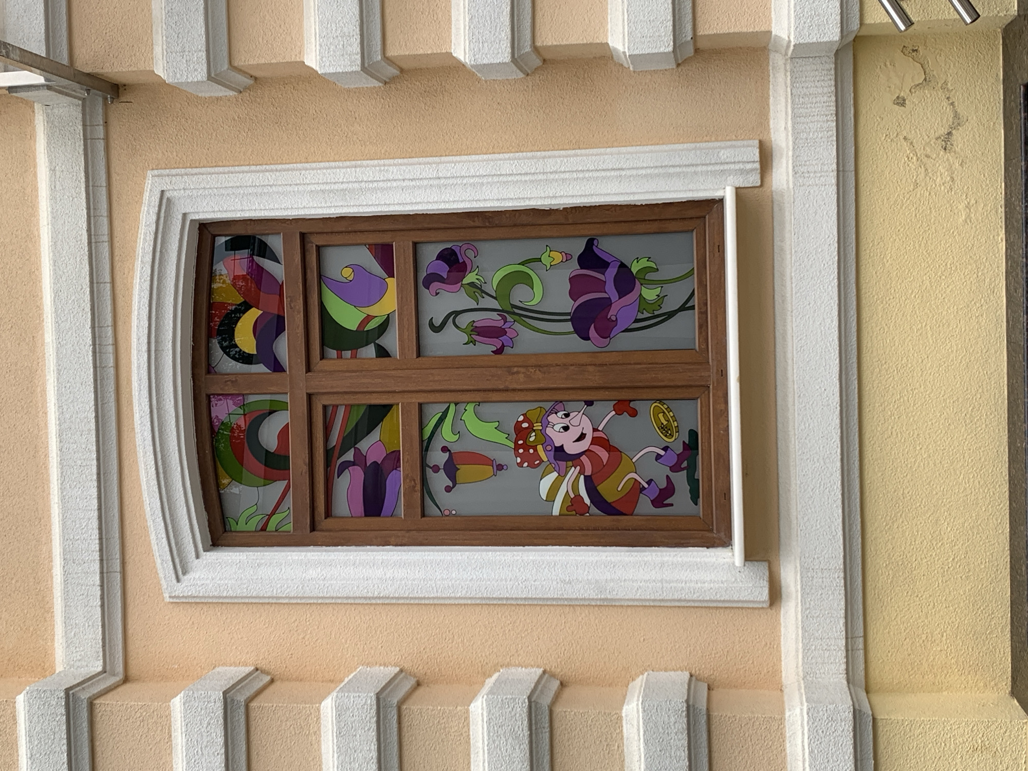 Here is a description of the image:
The image presents a close-up of a decorative window installed in a building exterior in Stavropol, Russia. The window is framed by a wooden border and divided into six panes. Each pane contains colorful, cartoon-like images of flowers and a whimsical insect-like character holding a coin. The window is set into a wall painted in a light peach color. The wall is bordered by white decorative molding above and below the window, as well as to the left. To the right of the window, the wall is a slightly different shade of yellow and shows signs of peeling paint or damage. The architectural style of the building includes ornate details, evidenced by the decorative molding and contrasting colors.