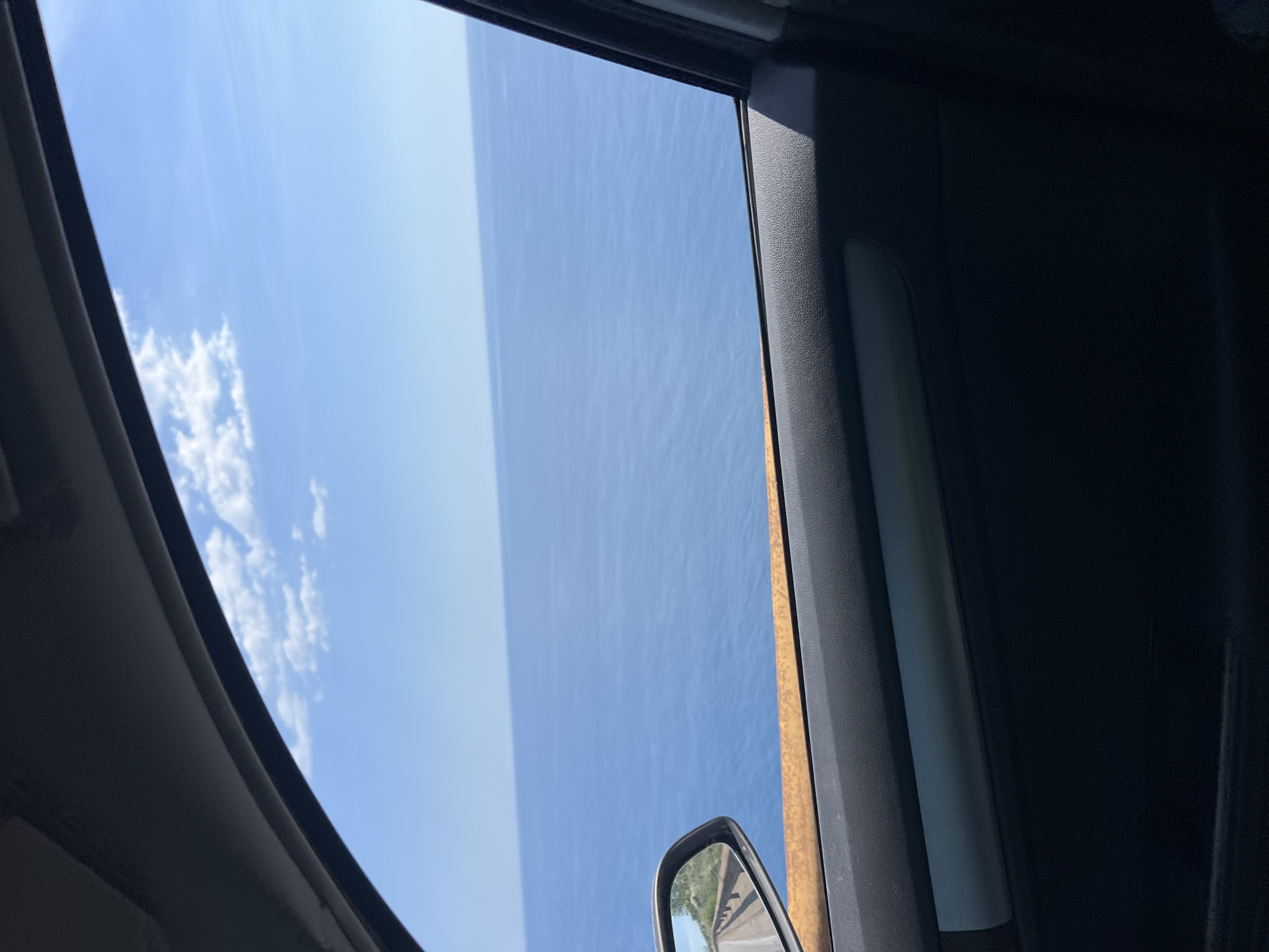 The image captures a view from inside a vehicle looking out of a partially opened window towards a vast expanse of the Adriatic Sea on a clear, sunny day. The water is a deep blue, meeting a pale blue sky dotted with sparse white clouds. In the side mirror, a road and a rust-colored guardrail are visible, suggesting the vehicle is traveling along a coastal route. The interior of the car, including the door panel and a reflective strip, is also visible in the foreground, framing the scenic ocean view. The location is identified as Dobra Voda, Montenegro.