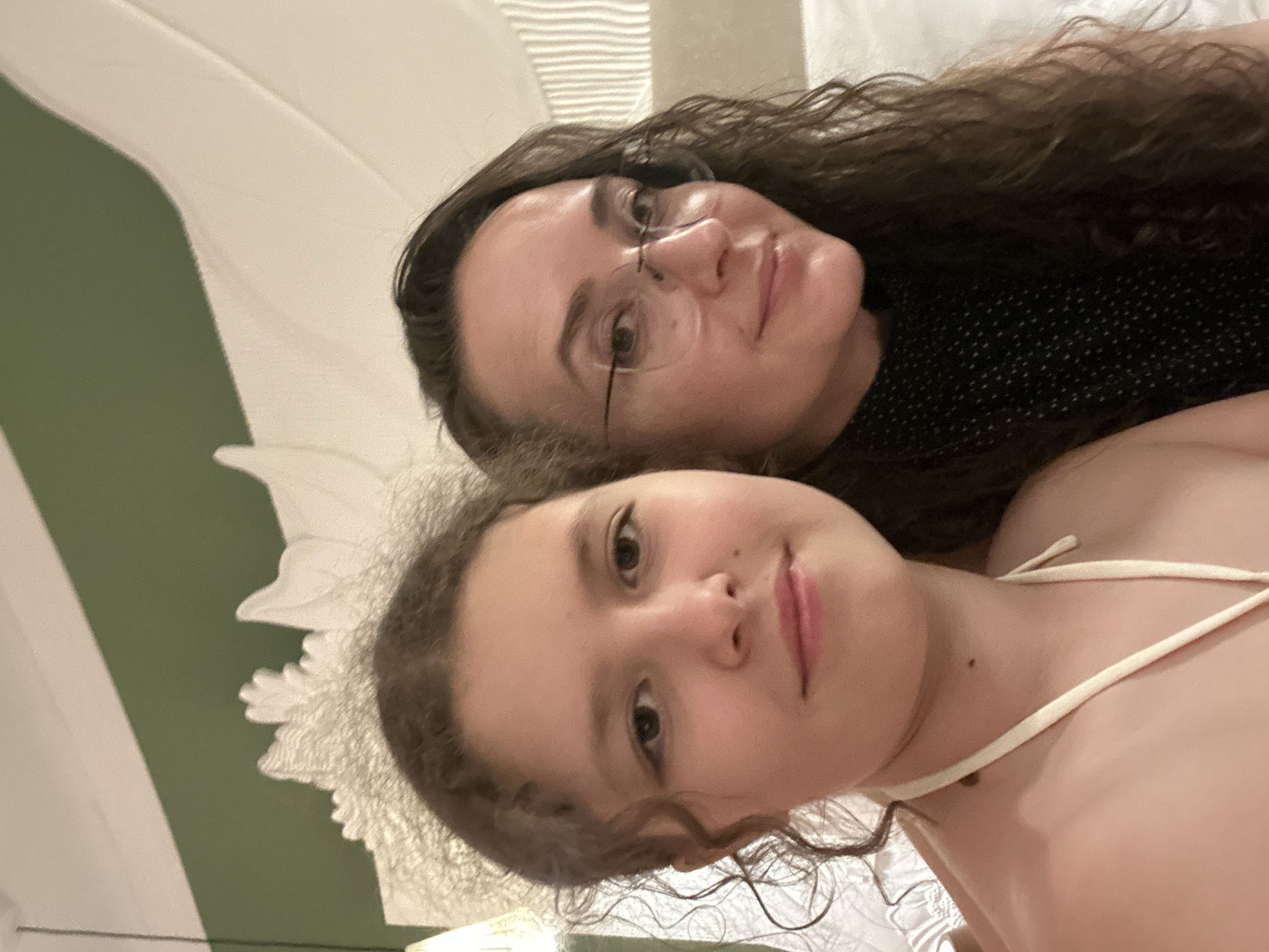 Two individuals, a younger female and an adult female, are posing for a selfie. The younger female is on the left, with curly brown hair and fair skin, wearing a white halter top. The adult female is on the right, with long, wavy dark brown hair, wearing glasses and a black patterned top. They are indoors, with a green wall and a white draped canopy visible in the background. The overall lighting suggests an indoor setting, possibly a hotel room or resort. This location is in Halaveli, Maldives.