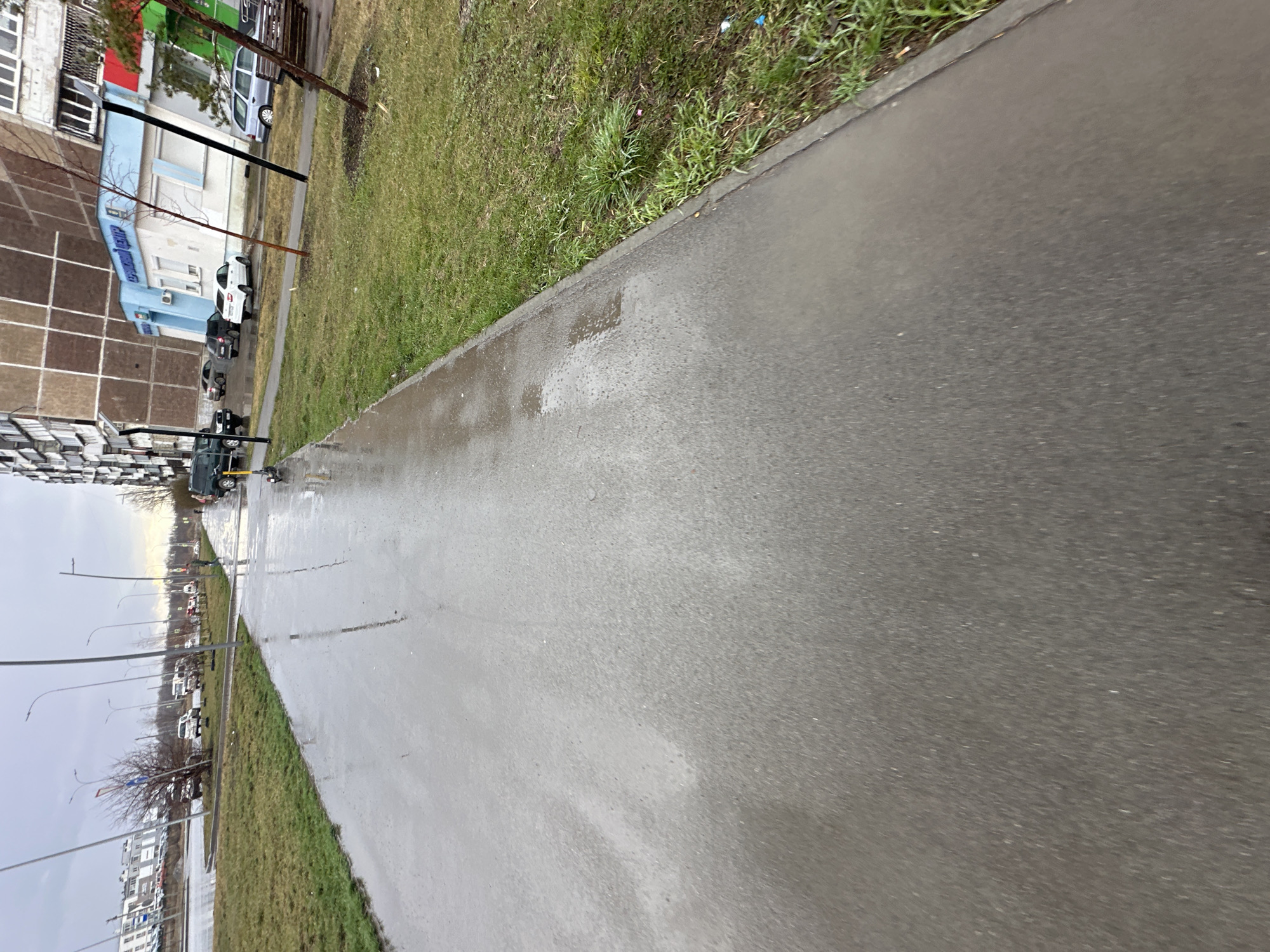 The image shows a wet sidewalk and road in an urban environment. The location appears to be in Unknown, Unknown. On the left side of the image, a building with a brown and white facade is visible. Adjacent to the building is a patch of green grass. Cars are parked along the side of the road, and streetlights line the street. The sky is overcast, contributing to the wet and somewhat dreary atmosphere.