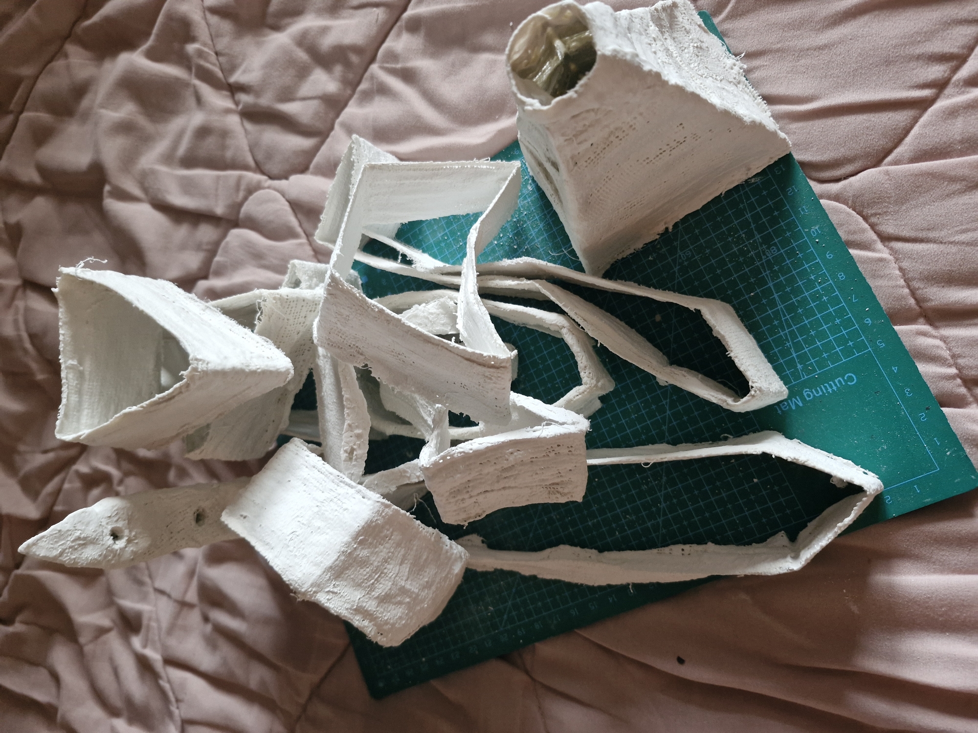 The image shows a collection of white, geometric, possibly plaster-like objects arranged on a green cutting mat, which rests on a pinkish-brown, textured surface, likely a bedspread or similar fabric. The objects appear to be fragments or components of a larger structure or model, featuring sharp angles and open spaces. Some have a cup-like shape, while others resemble strips or pieces of wall. The green cutting mat is marked with a grid pattern and numerical markings along its edges. The background surface is soft and rumpled. It is not possible to determine the location in Sofia, Bulgaria, from this image.
