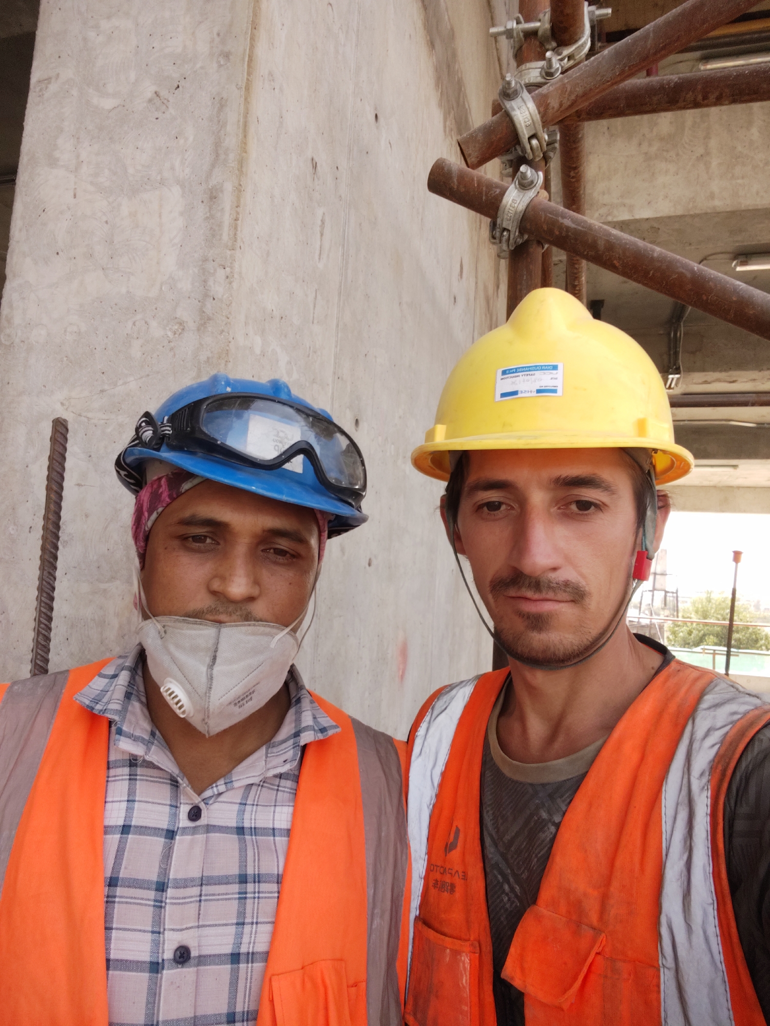 Two construction workers are taking a selfie outdoors. The worker on the left is wearing a blue hard hat with goggles, a pink bandana, a white respirator mask, a plaid shirt, and an orange safety vest. The worker on the right is wearing a yellow hard hat, an orange safety vest over a dark t-shirt. They are standing in front of a concrete wall and scaffolding. The background shows some trees and buildings, suggesting an urban environment. There is a rebar rod to the left of the man on the left. The scene appears to be a construction site. The image may have been taken in Dushanbe, Tajikistan, though this cannot be definitively confirmed from the image alone.