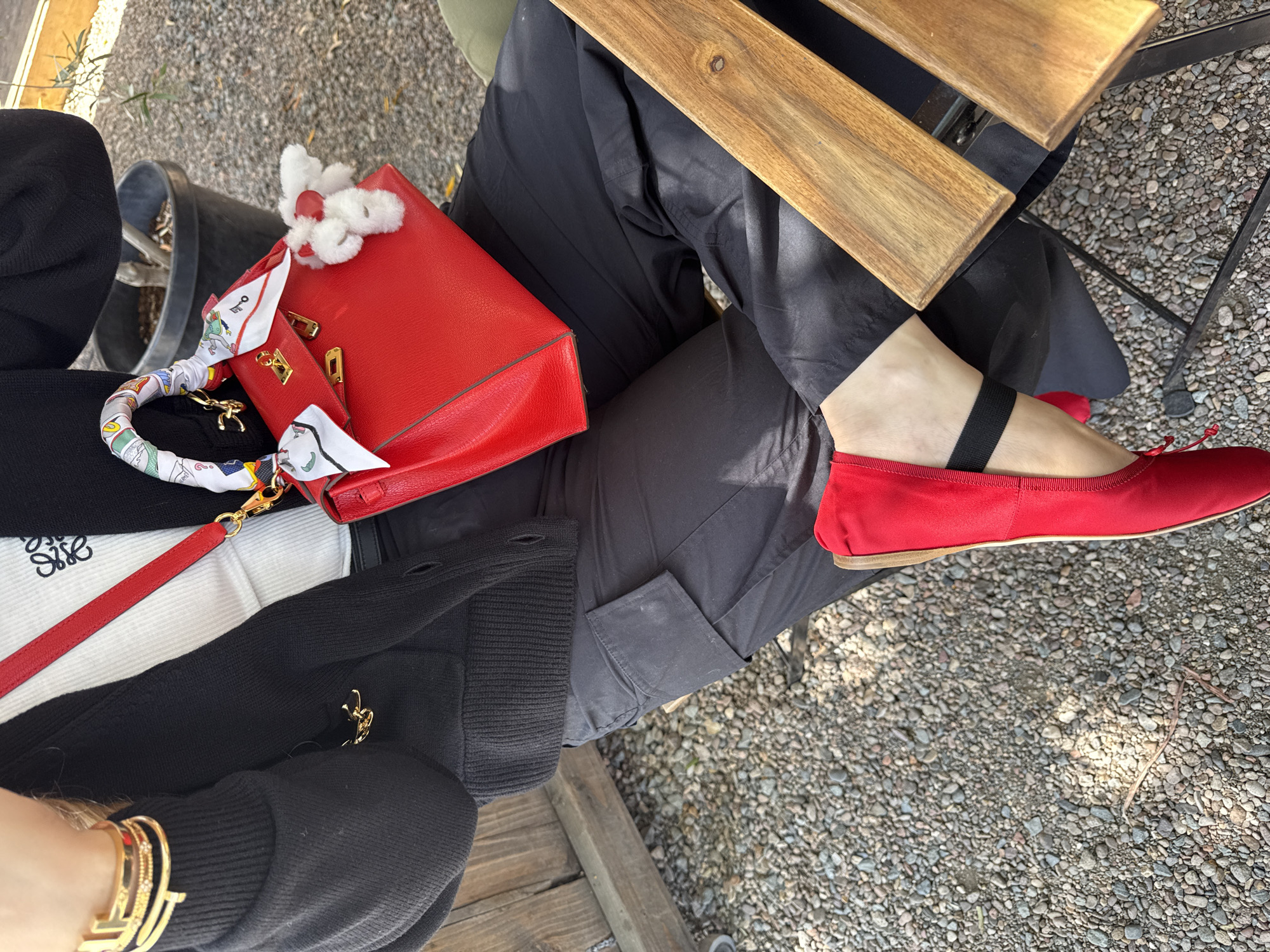 The image shows a person seated at an outdoor table on a gravel-covered surface. The person is wearing dark pants and red ballet flats with black straps. On their lap sits a red handbag with gold hardware, adorned with a white scarf and a small, white, fuzzy decoration. Part of a black and white top or sweater is visible, featuring a Loewe logo. A gold bracelet is visible on the person's wrist. A wooden table partially obscures the view of the person's legs. There is a plant pot in the background. The image suggests an outdoor setting, possibly a patio or garden. The specific location within the city of Chula Vista is not discernible from the image.