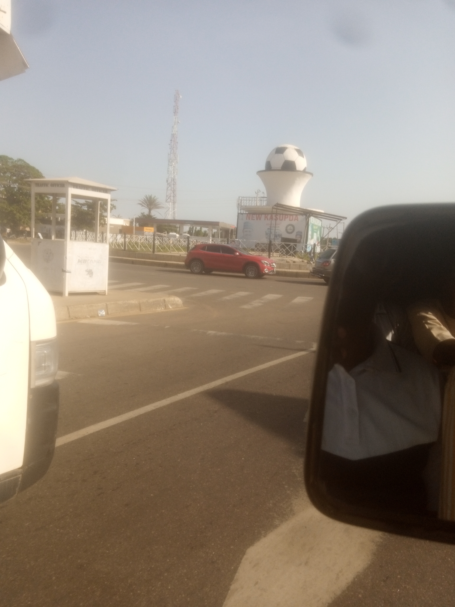The image shows a street scene in Sardauna, Nigeria, with a prominent roadside structure topped by a large white object resembling a soccer ball. A red car is visible on the road. In the foreground, a white vehicle is partially captured, and the reflection in its side mirror shows a portion of the interior and passengers. Another vehicle, a dark SUV, is parked behind the red car. A tall communication tower is visible in the distance. The environment appears to be a clear, possibly sunny day.