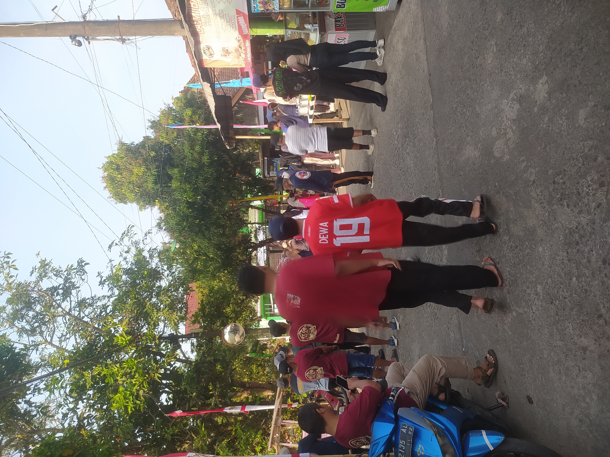 The image depicts a street scene in Lopait, Indonesia, with several individuals visible. In the foreground, on the left, a person is seated on a blue motorcycle, looking at their phone. To the right of the motorcycle, two people are walking away from the camera, one in black pants and the other in black pants with a red stripe. A person in a red jersey with the name "DEWA" and the number "19" is also visible, facing away from the camera.
In the middle ground, a group of people are gathered. Some are wearing matching red t-shirts with a circular logo that says "RT.OB". Other individuals are seen in various casual attire, including t-shirts, shorts, and pants.
The background shows a street lined with trees and buildings. A sign with text, including "LA MIDA" and "Fish & De", is visible on one of the buildings, suggesting a food establishment. Several flags, including Indonesian flags, are displayed. The sky is clear and bright, indicating daytime. The overall atmosphere appears to be a casual gathering or event.