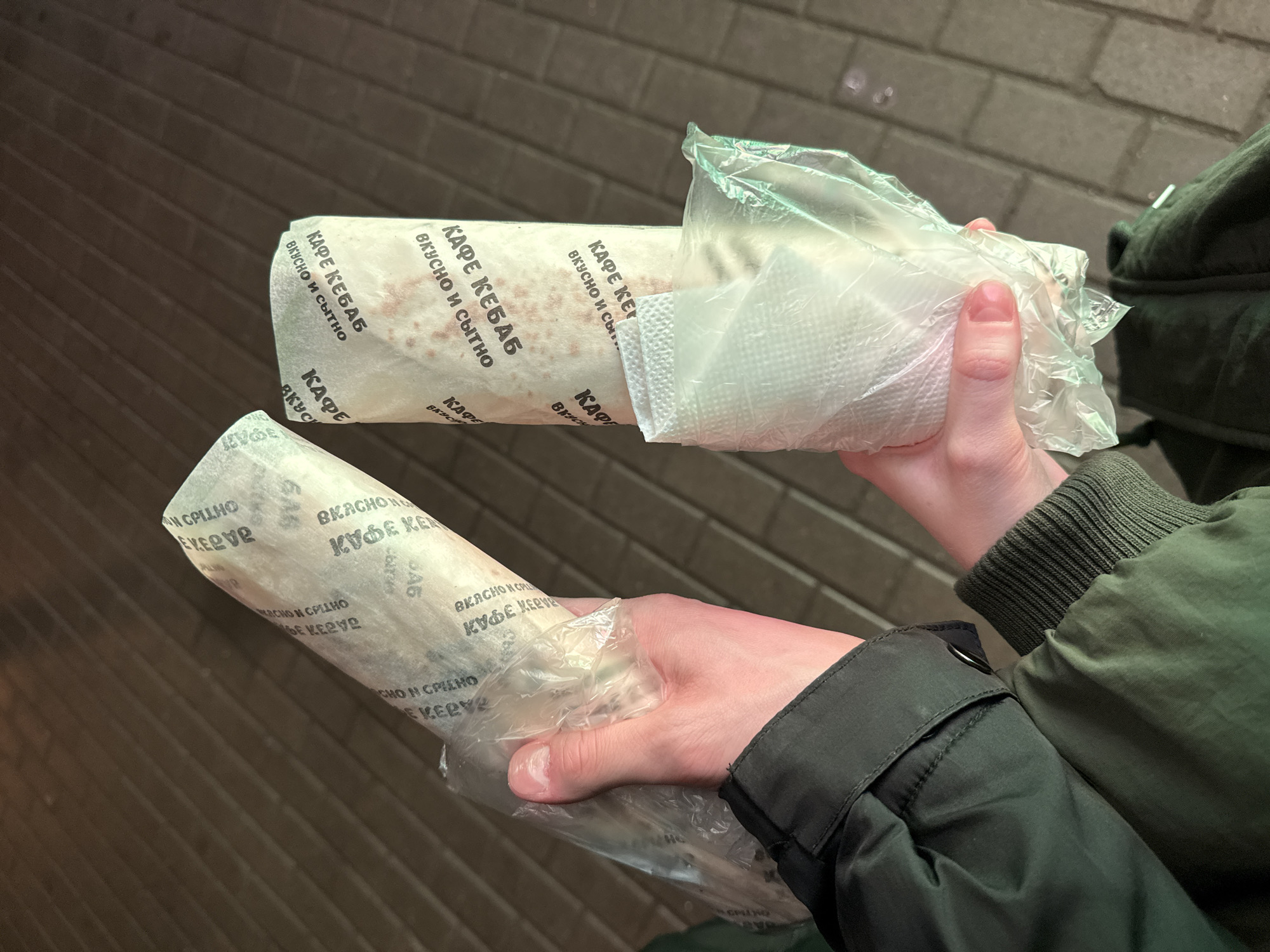 The image captures a first-person perspective of a person holding two large, wrapped food items, likely kebabs or shawarmas, in their hands, possibly in Saint Petersburg, Russia. The background shows dark, textured pavement or bricks, suggesting an outdoor urban setting, possibly a street or sidewalk, under dim lighting, indicating evening or night.
The hands, wearing parts of a dark green jacket with ribbed cuffs, hold the two food items. The item in the left hand is held firmly, while the item in the right hand is partially unwrapped, showing a clear plastic bag containing a white paper napkin over the food wrapper.
The paper wrappers of the food items are adorned with repeating text in Cyrillic script. The most prominent and legible text reads "КАФЕ КЕБАБ" (Cafe Kebab) and "ВКУСНО И СЫТНО" (Tasty and Filling), suggesting the origin and quality of the food. The repeated text is printed in a dark, bold font. The overall impression is of a casual, on-the-go meal, characteristic of street food.