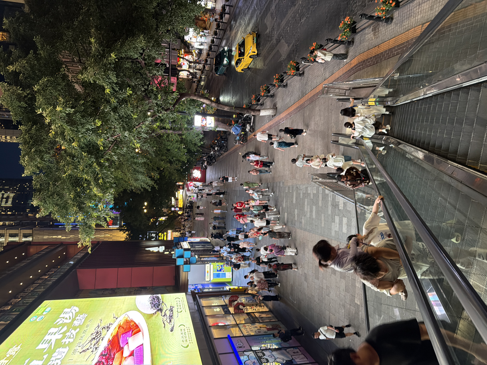 The image captures a nighttime scene on a bustling pedestrian street in Yuzhong District, China. A large illuminated billboard advertising food is prominently displayed on the left. The street is paved with textured tiles and lined with trees and decorative planters. On the right side of the street, vehicles, including a yellow taxi and a dark green car, are navigating a wet road. A diverse group of people are walking along the sidewalk and on an escalator. Among them, a child is being carried on another person's shoulders. The atmosphere is lively, with shop lights and streetlights illuminating the area.