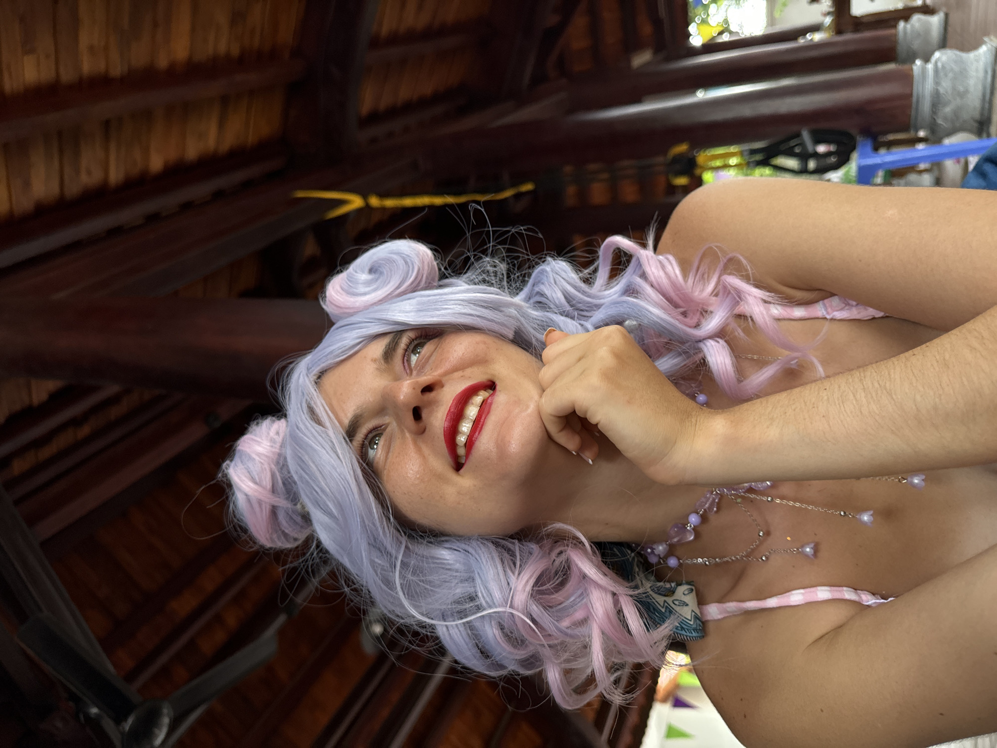 A woman with light skin and freckles, wearing a lavender and pink wig styled in two buns and loose curls, looks up and to the right with a smile. She has red lipstick on and is resting her chin on her right hand, with her left arm bent across her chest. She wears a pink and white checkered bikini top and a multi-layered necklace with purple beads and charms. The background shows the underside of a dark, wooden, sloped roof supported by thick wooden beams, with a ceiling fan visible in the upper left corner. The setting appears to be an outdoor covered area, possibly a restaurant or pavilion, with hints of greenery and other structures visible in the blurred background. The overall impression is a playful and whimsical portrait. The image appears to have been taken in Mũi Né, Vietnam, a coastal resort town known for its beaches, sand dunes, and laid-back atmosphere.
