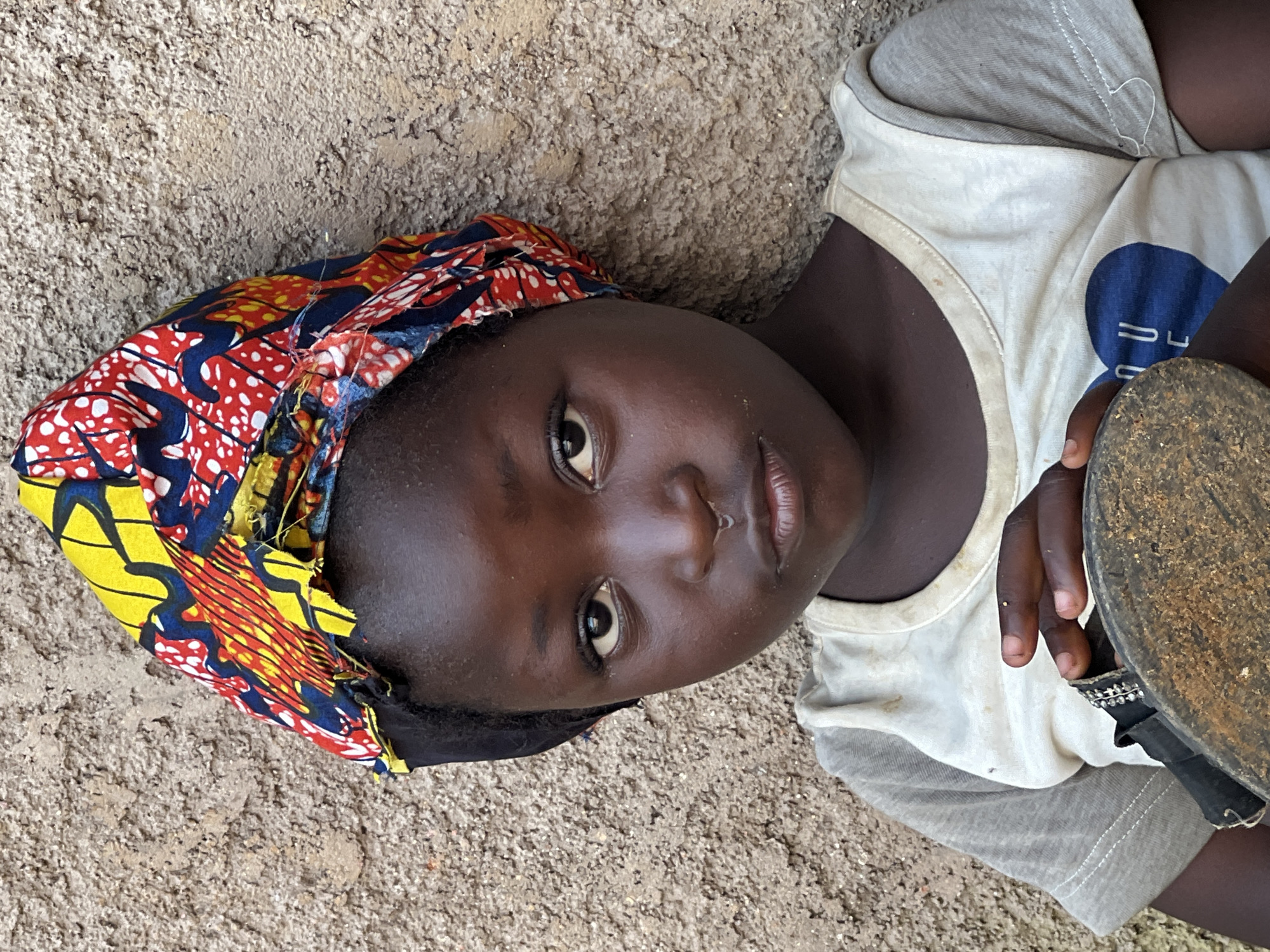 A young Black girl is shown in a close-up portrait against a rough, textured wall. She is wearing a colorful headscarf with a pattern of red, yellow, and blue, tied over a black under-scarf. Her t-shirt is light-colored with grey sleeves and features a blue heart graphic on the front. The girl is holding a worn, round object, possibly a sandal sole, in her hands. A faint drip is visible on her upper lip. The background wall appears to be made of concrete or stucco, showing signs of wear and tear. The lighting suggests it is daytime, possibly outdoors, in a location like Jos, Nigeria.
